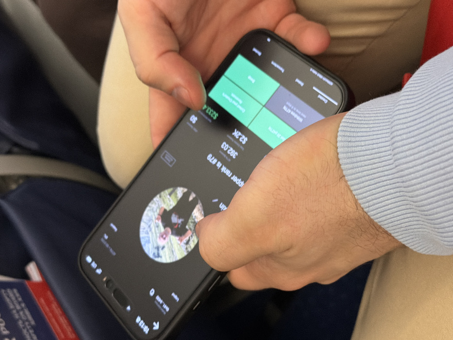 A close-up, slightly overhead image captures an individual holding a black smartphone with both hands, positioned within an aircraft cabin. The smartphone displays an active application with a dark interface. Centered on the screen is a circular profile image depicting a person in a dark t-shirt and head covering, making a peace sign gesture against an outdoor backdrop. Visible text below the profile includes "hopper rank is #79," "392.03," and "$2.2K." The right side of the screen features green panels with "$233.4" and options such as "Transfer Funds" and "Withdraw ATM," indicative of a financial or cryptocurrency-related application. The right thumb is positioned over the screen for interaction. The left wrist is covered by a light blue, ribbed sleeve. The background consists of blurred dark blue airplane seats and a partially visible red and white safety card with Russian text ("РОССИИ") inserted into a seat-back pocket. A light beige object, possibly a headrest or cushion, is also discernible. The scene is located within an airplane at or en route to Yemelyanovo International Airport (Емельяново (аэропорт)), Russia.