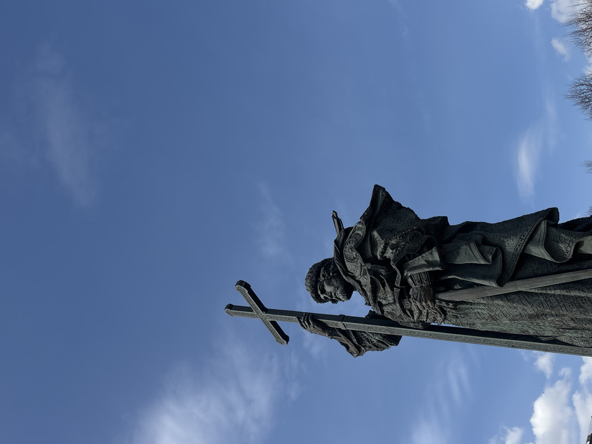 The image depicts a low-angle shot of the monument to Patriarch Hermogenes in Moscow, Russia. The bronze statue features a religious figure in Orthodox vestments, holding a cross in his left hand. The statue is silhouetted against a clear, blue sky with a few scattered clouds. Bare tree branches are visible in the upper right corner, indicating the season is likely late winter or early spring.