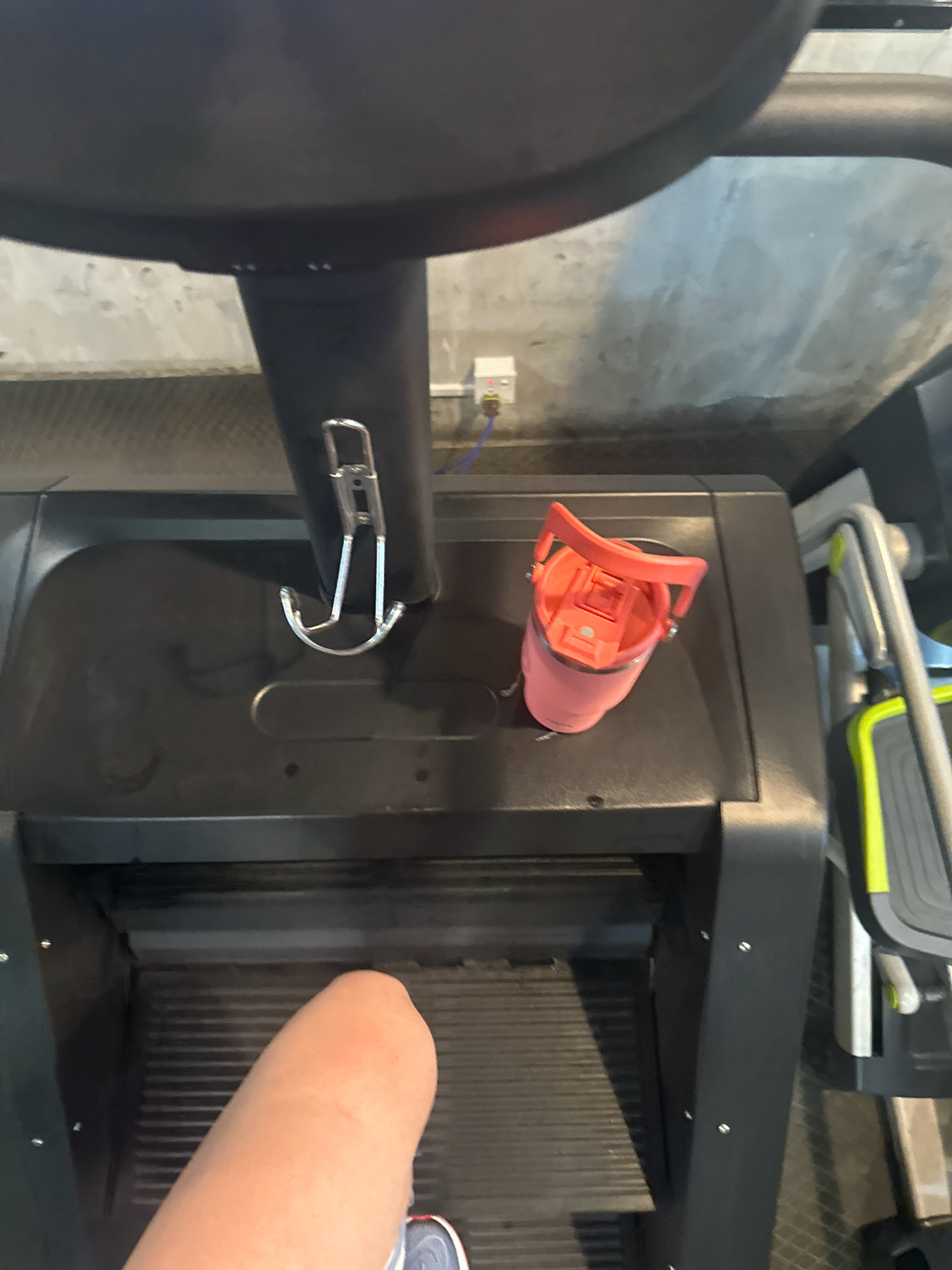The image shows a close-up, overhead view of a treadmill in a gym. A person's leg, from the knee down, is visible on the left side of the treadmill belt. A pink water bottle with a black lid and a red handle is placed on the right side of the treadmill's console. A metal hook is attached to the treadmill's console. In the background, a concrete wall is visible, with a white electrical outlet and some wires. Part of another piece of gym equipment, possibly an elliptical machine, is visible on the far right. The scene is well-lit, with a slight sheen on the treadmill surface and the concrete wall. The context of the image is a gym environment, suggesting a workout is in progress or has recently concluded. There is no identifiable information in the image that would indicate its location in Lalitpur, Nepal.