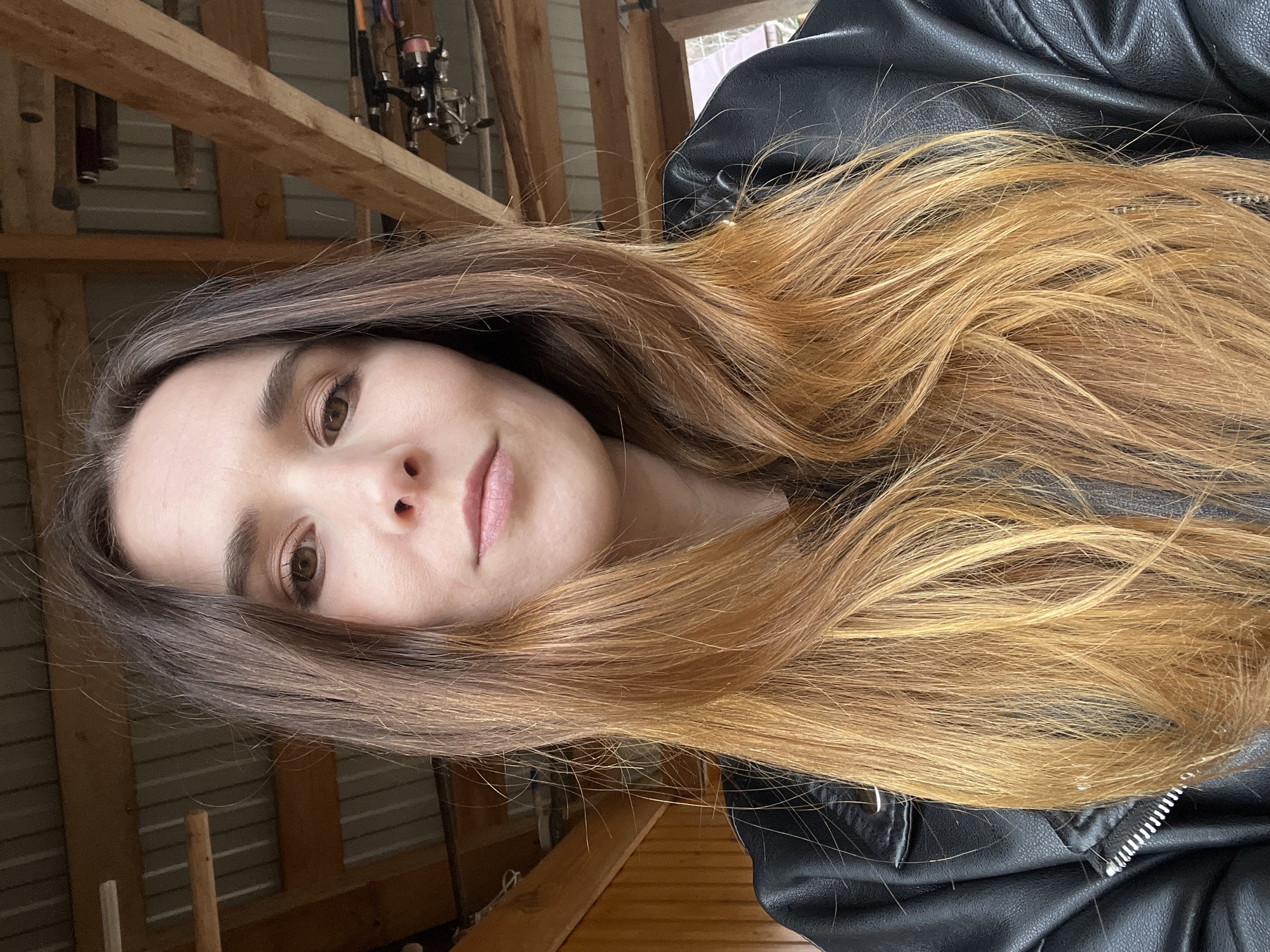 The image is a close-up selfie of a young woman with long, ombre hair transitioning from dark brown at the roots to golden blonde at the tips. She has fair skin, brown eyes, and is wearing light pink lipstick. She is wearing a black leather jacket. The background is blurred and out of focus, but appears to show the interior of a wooden structure with exposed beams and metal sheeting. Fishing rods and reels are visible hanging in the background. The location in the city of Kamskoe, Russia is not discernible from the image.