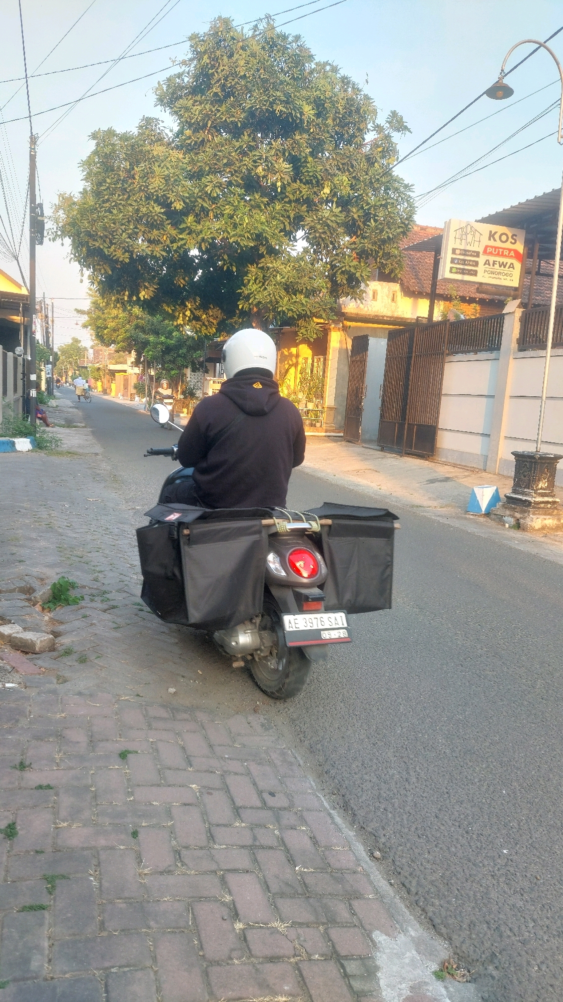 The image depicts a man wearing a white helmet and a black hoodie riding a scooter down a paved street in Kertosari, Indonesia. The scooter is equipped with two large black saddlebags on either side of the rear. A license plate on the scooter reads "AE 3976 SAI". The street is lined with a brick sidewalk on the left and buildings and fences on the right. A large tree with green leaves stands prominently in the background. The sky is clear and appears to be early morning or late afternoon, with the sun casting long shadows. A sign for "KOS PUTRA AFWA" is visible on the right side of the street.