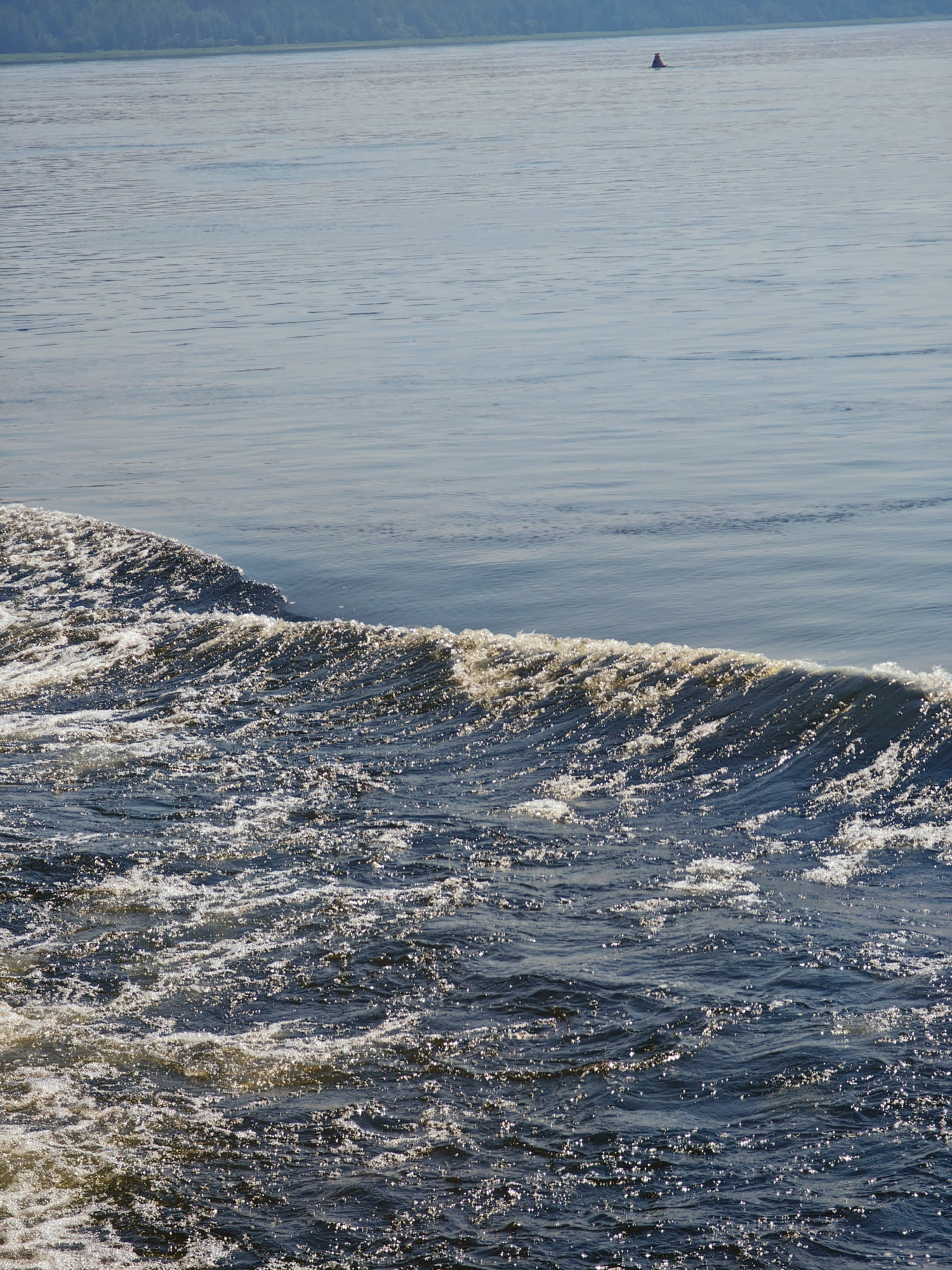 The image depicts a body of water, likely a river, in Pogodaevo, Russia. The foreground is dominated by choppy, dark blue water with sunlight glinting off the surface, creating a foamy, turbulent wake. A distinct wave crests from left to right across the mid-section of the image. The background shows calmer, lighter blue water stretching towards a distant, tree-lined shore. A small red and white buoy is visible in the upper right quadrant of the scene, floating on the calmer water. The overall scene is natural, with no people or man-made structures immediately apparent, apart from the buoy.