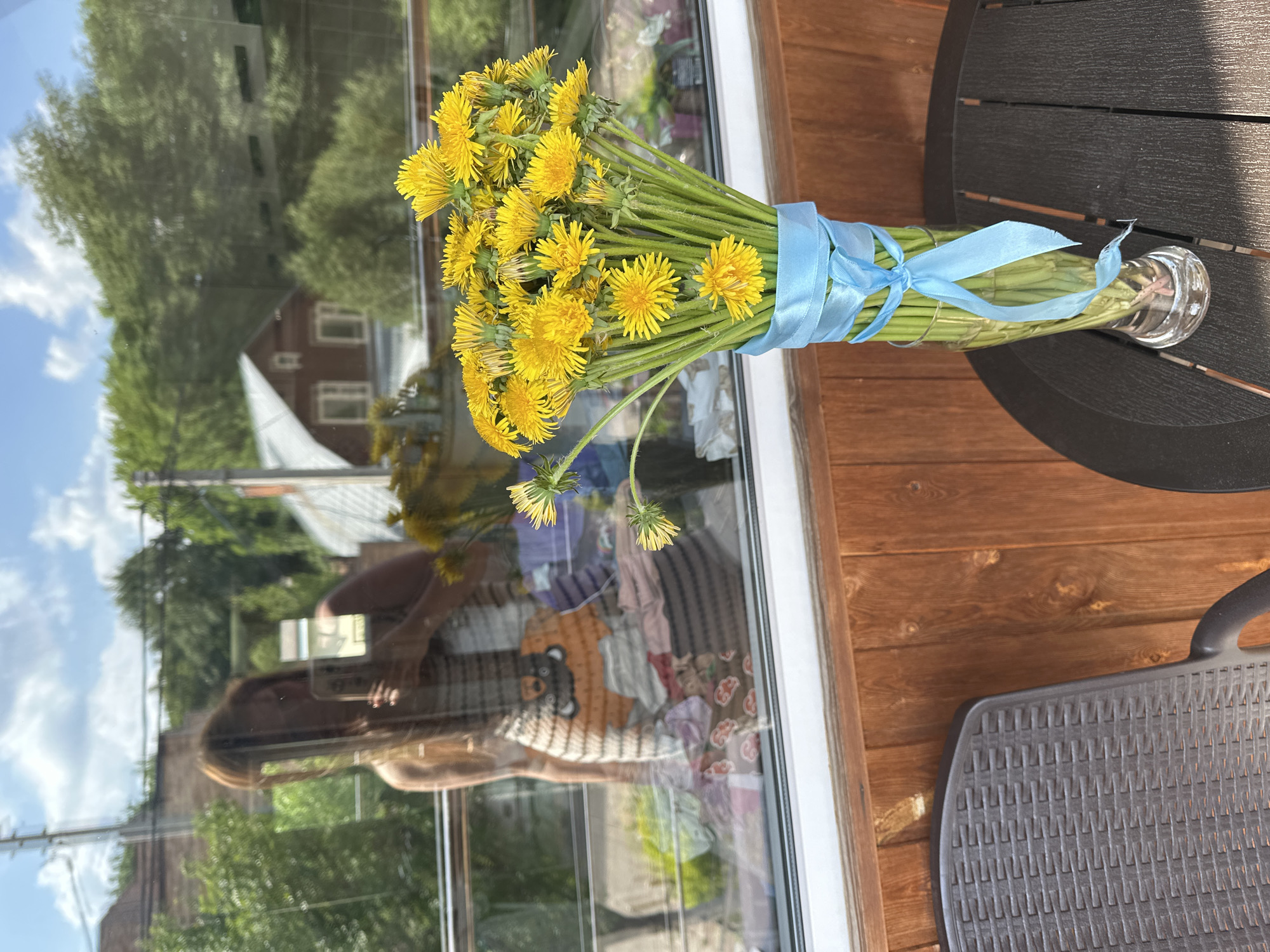 A still life photograph depicts a bouquet of dandelions secured with a light blue ribbon in a clear glass vase, positioned on a wooden surface adjacent to a black patio table. The bouquet is primarily composed of yellow dandelion flowers with green stems. The background showcases a reflection in a window, revealing a partial view of a house with light-colored siding and a white roof, along with greenery and a blurred human reflection. The scene is captured in daylight with natural lighting. The image is taken in Nizhny Novgorod, Russia.