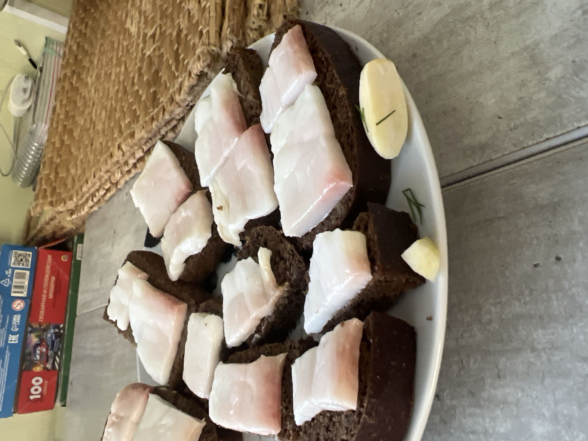 The image shows a plate of dark bread slices topped with slices of pork fat. A small piece of cheese and a sprig of dill are also on the plate. The plate is on a wooden table. In the background, there are some boxes, a woven mat, and a wall. The location is not specified in Volgograd, Russia.
