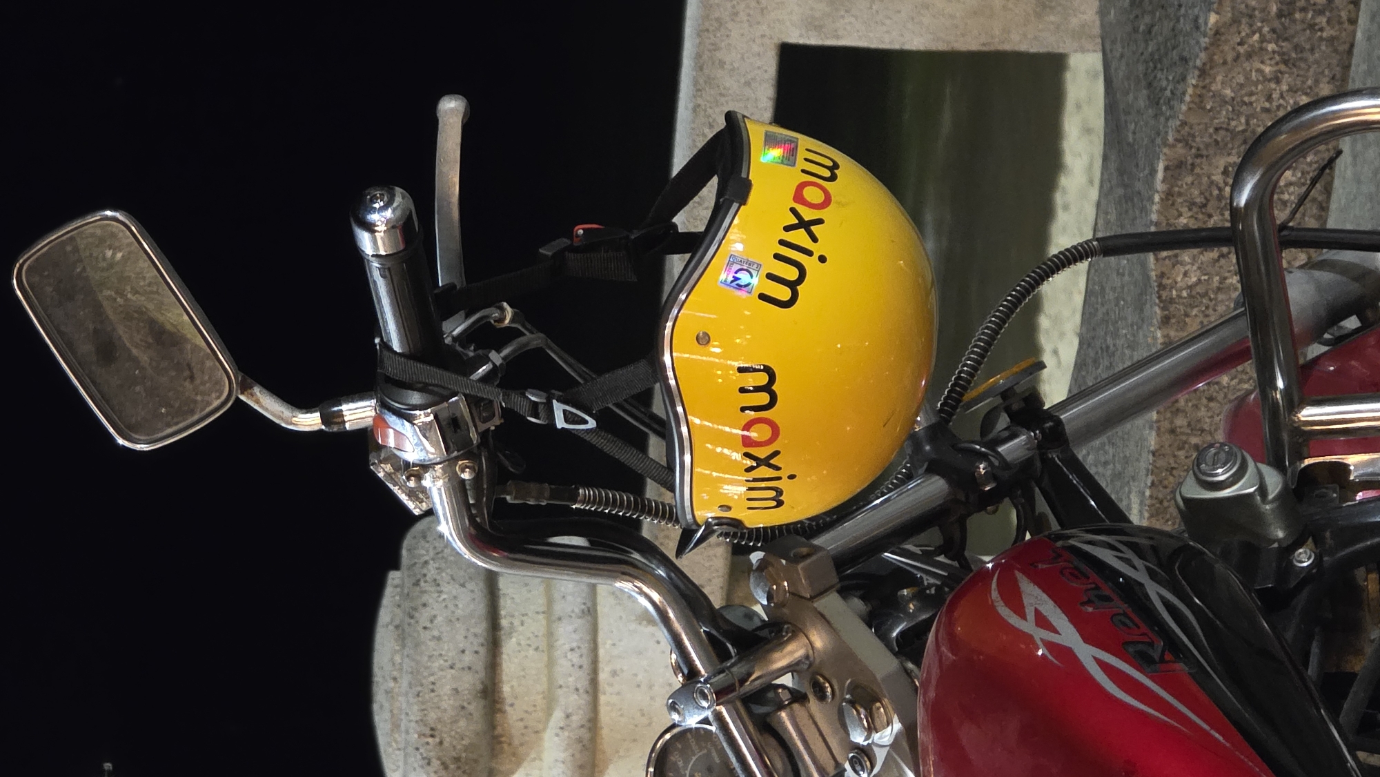 The image captures a close-up of a parked motorcycle with a yellow helmet hanging on its handlebars, set against a dark background, suggesting nighttime or low-light conditions. The motorcycle features chrome handlebars, a rectangular rearview mirror on the left, and visible cables and controls. Its fuel tank is predominantly red with black accents and intricate silver or white tribal-style graphics that clearly spell out "Rebel".
The bright yellow motorcycle helmet is prominently displayed, secured by its strap to the handlebars. The helmet has the word "maxim" printed multiple times in black text, oriented vertically, indicating it belongs to a driver for the Maxim ride-hailing service. A small holographic sticker and another sticker bearing the text "QUATEST 3" are also visible on the helmet. Reflected light highlights the chrome surfaces of the motorcycle and the glossy finish of the helmet. A light-colored, possibly concrete or stone, pillar or wall is partially visible in the blurred background.
The scene depicts a stationary vehicle, with no human subjects. Given the context of Nha Trang, Vietnam, the "Maxim" branding on the helmet is a common sight for ride-hailing and delivery services in the region. The overall impression is a candid shot of a motorcycle and rider's equipment, likely awaiting a passenger or parked after a ride.