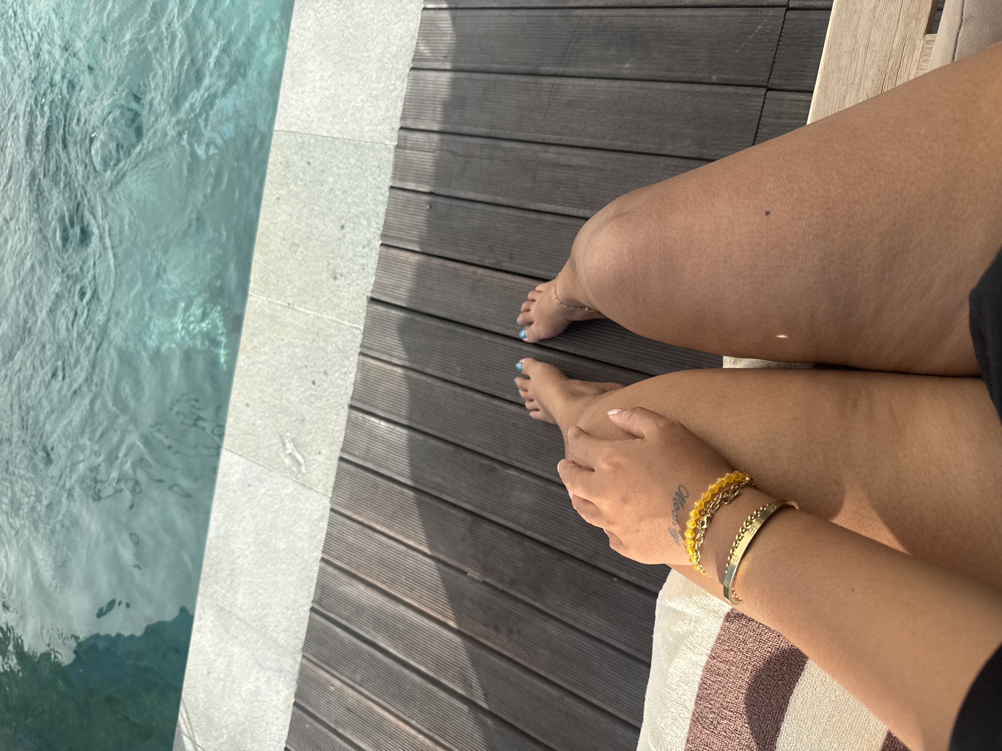 A high-angle shot captures a person's legs and arms resting on a lounge chair next to a swimming pool. The pool's turquoise water is visible in the upper portion of the frame, bordered by a light stone ledge and dark wooden decking. The person's skin is tanned, and they are wearing blue nail polish on their toes. Their left arm is adorned with two gold bracelets, one with yellow beads. A faint tattoo is visible on their left wrist. The setting appears to be a resort or private residence in North Kuta, Indonesia, a popular tourist destination known for its beaches and villas.