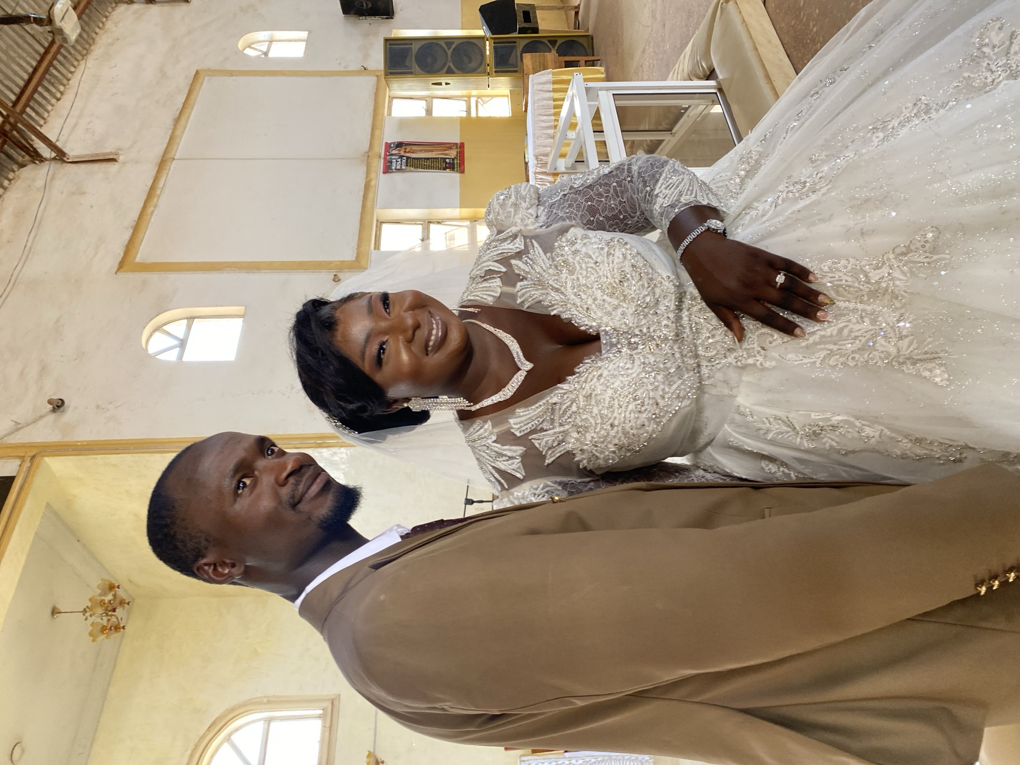 A wedding photograph taken inside a building in Kuru Karama, Nigeria, features a bride and groom. The bride, wearing an ornate white gown with a full skirt and long sleeves, a bejeweled necklace and bracelet, and a veil, smiles broadly at the camera. She is holding her left hand outward, displaying a wedding ring. The groom, wearing a light brown suit and white shirt, looks up toward the bride. The building has yellow walls, arched windows, and speakers are positioned high on the walls. A chandelier hangs from the ceiling.