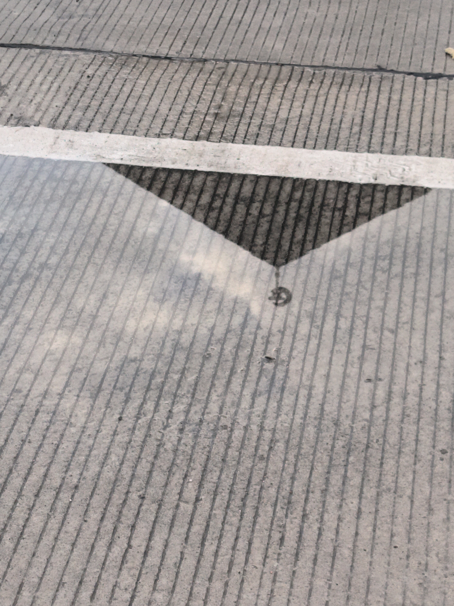 The image presents a ground-level view of a concrete surface, likely a road or paved area in Keureuweungkrueng, Indonesia. The surface is characterized by numerous parallel grooves running diagonally across the frame, providing texture and linearity. A prominent white line, painted horizontally, bisects the image roughly through its middle.
Below this white line, a puddle of water has formed, creating a reflective surface that covers the lower half of the photograph. Within this puddle, a dark, triangular shape is clearly reflected, resembling a shadow or a structural element above. The apex of this dark triangle points downwards into the puddle. The grooved texture of the concrete is also visible through the water's reflection, appearing lighter and somewhat blurred compared to the dry sections.
A notable detail within the reflection, near the pointed end of the dark triangle, is a small, faint circular symbol. This symbol appears to be a stylized letter 'G' or a simple logo, adding an intriguing element to the reflected scene.
The presence of the puddle indicates recent rainfall or wet conditions. The lighting suggests it is daytime. The scene is static, focusing on the interplay of concrete textures, painted lines, and reflections on a wet surface. No people or specific events are depicted.