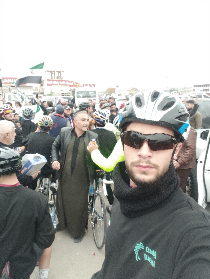 A selfie shows a man in cycling gear including a helmet, sunglasses, and neck gaiter. He wears a black sweatshirt with Arabic text. Behind him are several cyclists and pedestrians, many also wearing helmets. A man in a leather jacket and long, brown robe stands out. Bicycles are visible amongst the crowd. In the background, buildings and parked cars are seen with a flag displaying green, white, and black horizontal stripes. The scene seems to be an event or gathering in an urban environment on a cloudy day.