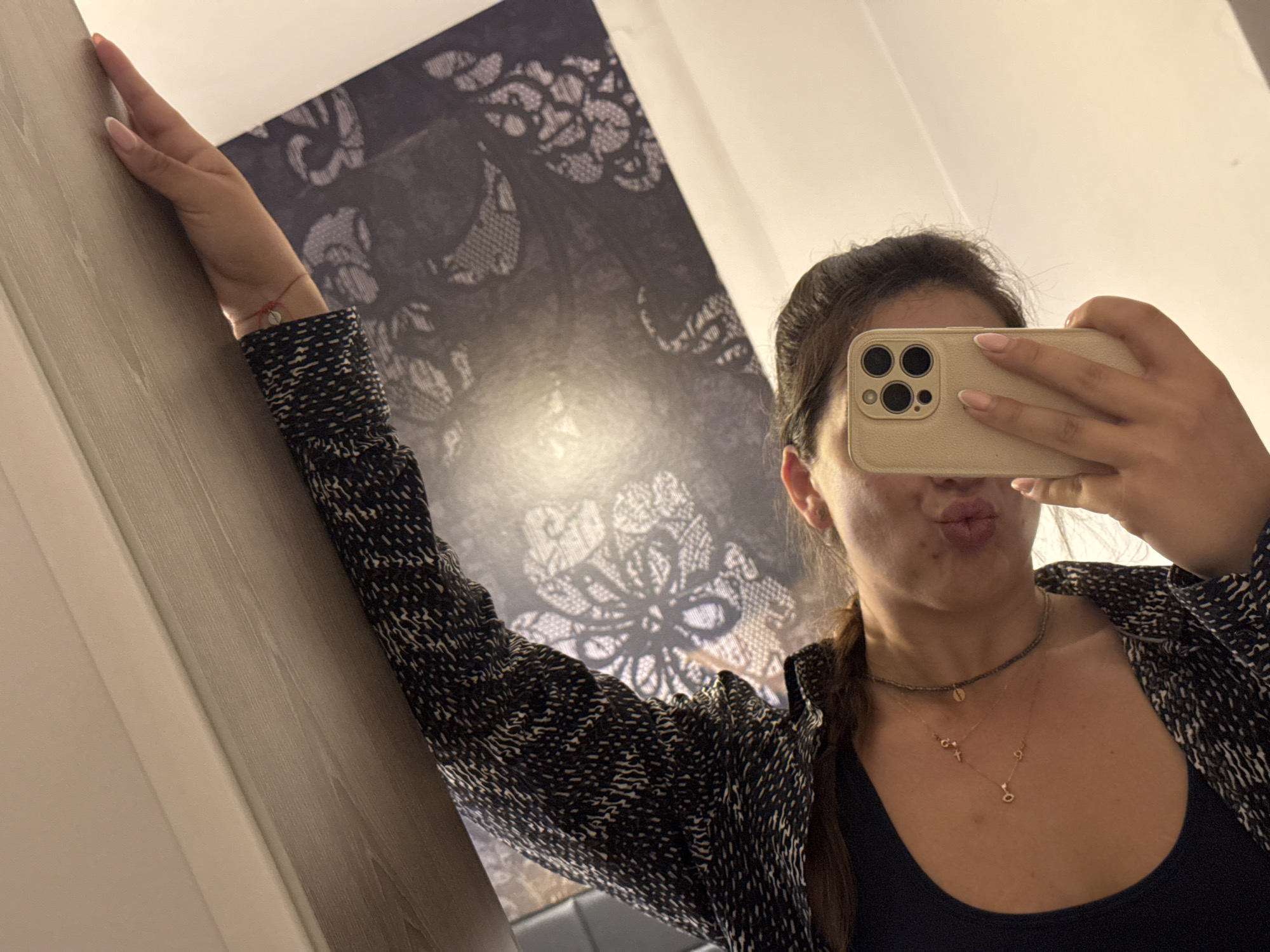 A young woman is captured taking a mirror selfie indoors. Her right arm is raised and bent, leaning against a light-colored wooden doorframe or wall panel on the left side of the frame. She holds a light-beige smartphone with a triple-lens camera in her left hand, capturing her reflection. Her lips are puckered in a playful expression, suggesting a kissy face.
The woman has dark hair, styled away from her face, with a section of hair, possibly a braid, visible over her left shoulder. She is dressed in a dark, intricately patterned long-sleeved top or jacket, worn over a plain dark tank top. She wears several necklaces: a dark, choker-style beaded necklace, and two delicate gold-colored chains with small pendants, one of which resembles the letter 'o' or a heart. A thin red string bracelet with a small silver charm is visible on her right wrist. Her fingernails are neatly manicured with a light, neutral polish.
The background features a large, dark wall or tapestry adorned with a prominent, ornate lace-like pattern in shades of dark gray or black. The upper right corner of the image shows a plain light-colored wall or ceiling. The indoor setting is illuminated by artificial light. No visible text is present in the image. This scene is located in Sofia, Bulgaria.