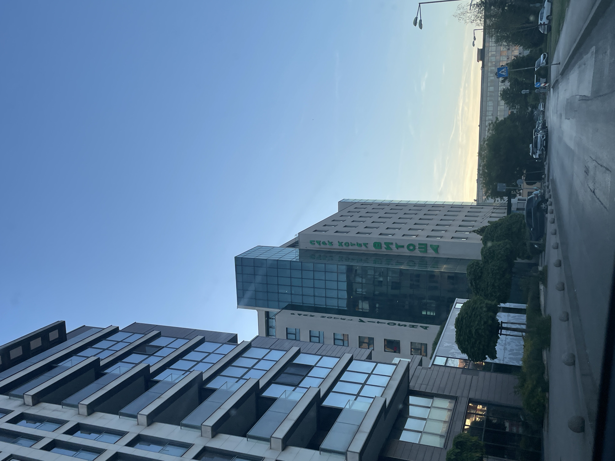 The image shows a street view in Sofia, Bulgaria, featuring modern architecture. Dominating the scene are two buildings: one with a distinctive pattern of slanted, overhanging architectural elements and another taller building with a glassy section and the name "VITOSHA" displayed in green, reflective lettering. The street is visible on the right side, with trees and parked cars. The sky is a clear blue, transitioning to a pale yellow near the horizon, suggesting either sunrise or sunset. The view is somewhat obstructed by a dark element, likely a car window.