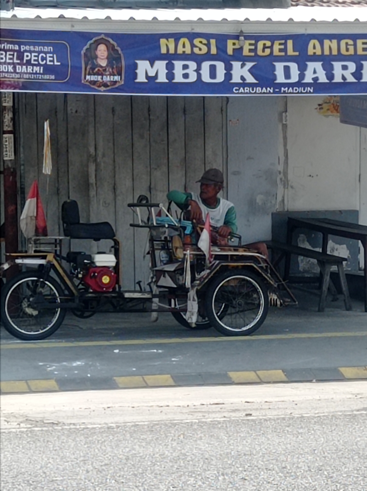 A man is seated on a motorized tricycle with a red engine. The tricycle has two wheels in the front and one in the rear. The man is wearing a brown cap and a white and green shirt. He is looking to the left. Behind him, a banner reads "NASI PECEL ANGE MBOK DARMI" with "CARUBAN - MADIUN" below it. The tricycle is parked on a street in what appears to be an outdoor market or roadside stall area, indicated by the wooden stall structures and the presence of a bench. The ground is paved, with yellow and black speed bumps visible. The sky is overcast.