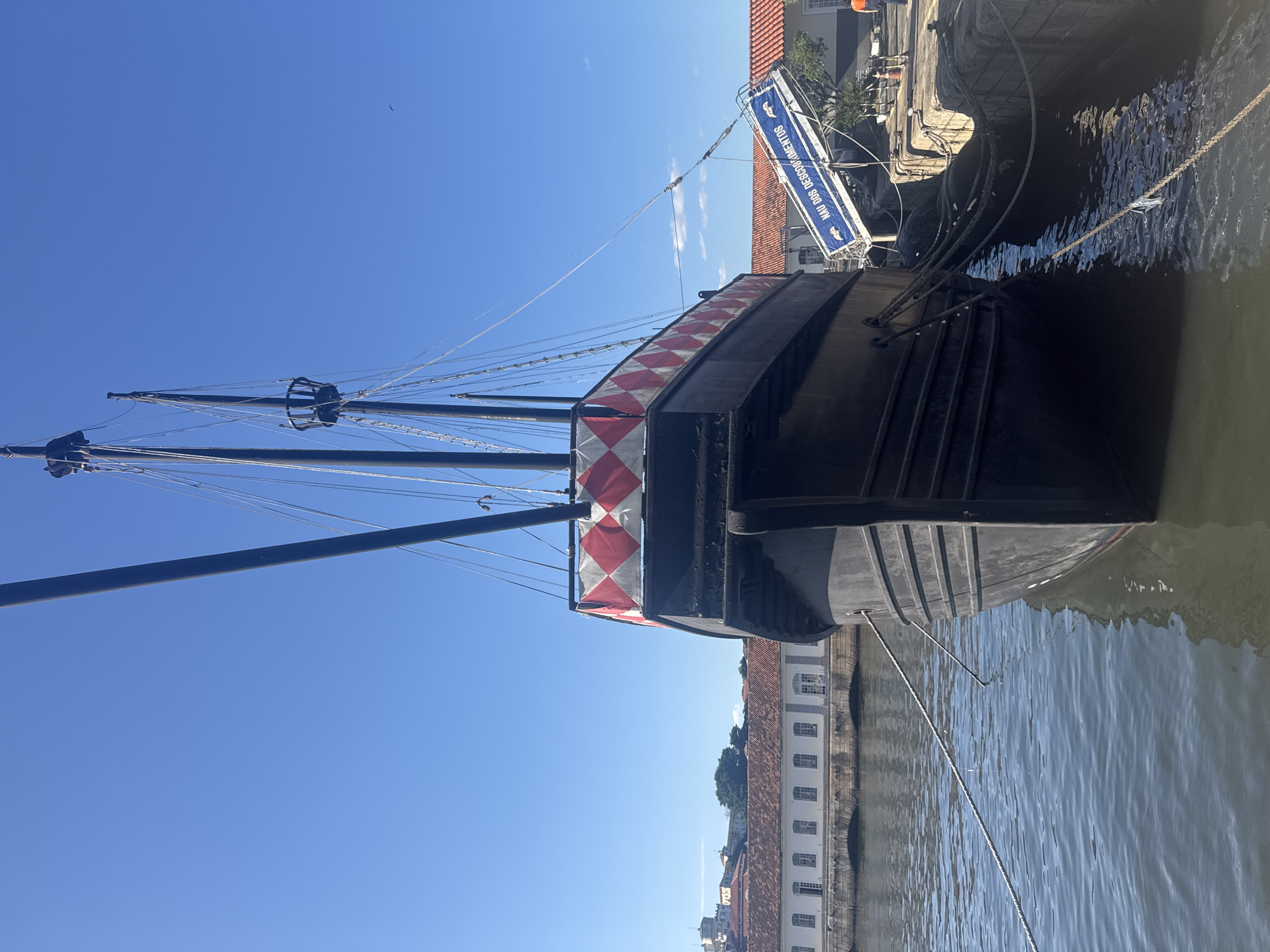 This is a low-angle, full-shot, eye-level photograph taken outdoors on a clear day. The main subject is a large, dark-colored replica of a historical sailing ship docked alongside a stone pier. The ship features tall masts with intricate rigging, and its hull is visible at the waterline, with thick ropes tethering it to the dock. The upper part of the ship's hull is adorned with a decorative band of red and white diamond-shaped patterns. A ramp labeled "NAU DOS DESCOBRIMENTOS" (Ship of Discoveries) extends from the dock to the ship, suggesting it is a tourist attraction.
In the background, across a body of water, is a long, low building with a red-tiled roof and numerous arched windows, characteristic of colonial architecture. Further in the distance, a hill with some vegetation and other buildings can be seen. The sky is a vibrant, cloudless blue. The water is calm, reflecting the ship and the sky. The scene appears to be located in the port area of Rio de Janeiro, Brazil, likely at the Praça dos Navegantes or a similar historical maritime site. No people are prominently featured in the foreground, though a person in an orange shirt is visible on the dock in the far right background. The overall atmosphere is peaceful and sunny.