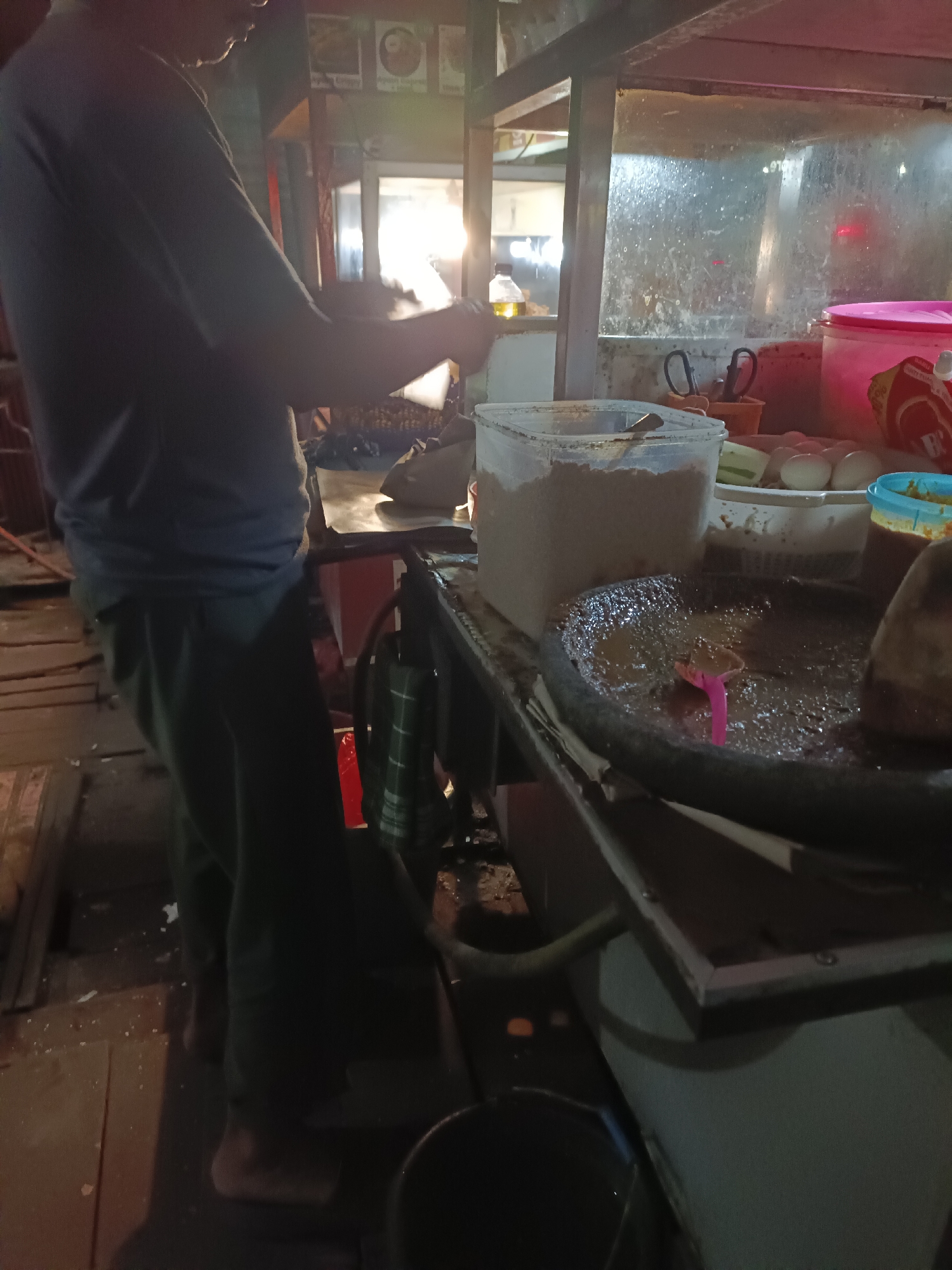 The image depicts a street food stall in Sangatta, Indonesia, at night. A person, partially visible from the side, is standing behind a stall that appears to be preparing food. The stall is equipped with various containers, one of which holds a light-colored powder. In the foreground, a stone mortar and pestle are visible, filled with a dark, textured mixture, suggesting the preparation of a paste or sauce. Several boiled eggs are arranged in a white bowl on a shelf above the mortar. The overall environment is dimly lit, with light emanating from the stall itself and possibly from the surrounding area. The stall structure includes metal framing and what appears to be a transparent panel. The ground beneath the stall is a mix of wooden planks and dark, possibly dirt or asphalt. A red bucket and a checkered cloth are also visible near the base of the stall.