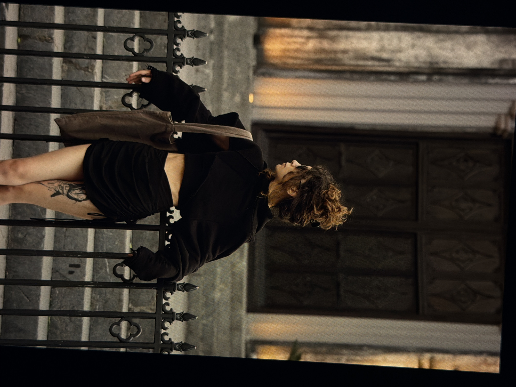 A sideways-oriented medium shot captures a woman leaning against a black wrought-iron gate. She is wearing a black outfit composed of a top and a skirt, and a light brown bag hangs over her shoulder. A tattoo is visible on her left leg. Her curly hair is pulled up in a bun.
Behind the gate are stone steps and a building with dark brown, ornate windows. The scene appears to be located outdoors in Đà Nẵng, Vietnam.
