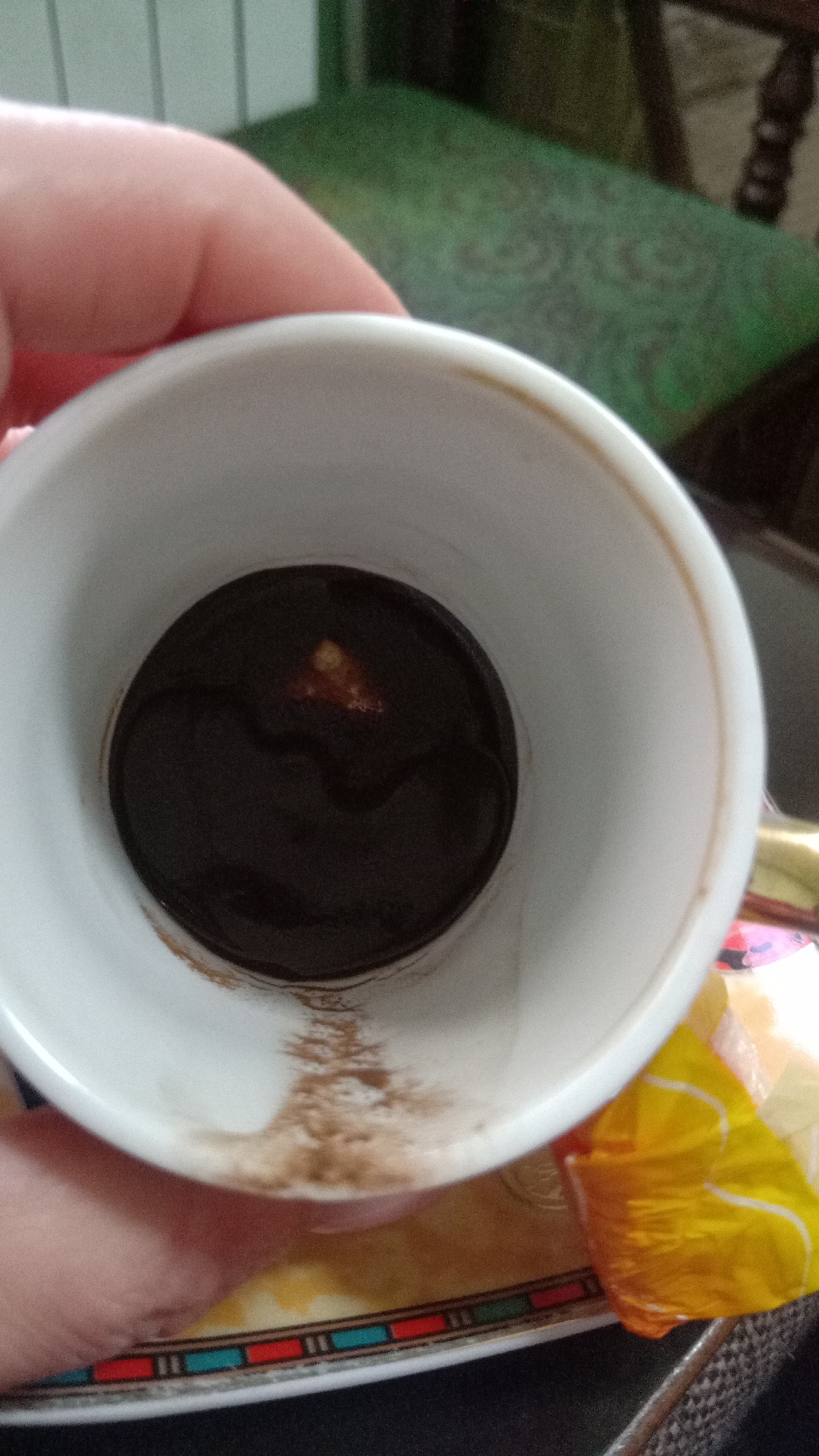 A close-up shot shows the interior of a white coffee cup with dark coffee residue at the bottom. A small, light-colored particle floats in the residue. Coffee stains streak down the inside of the cup. The cup sits on a colorful saucer, partially obscured by a piece of yellow and white plastic wrapping. A hand holds the cup. The background includes a green chair with an ornate pattern on the seat.