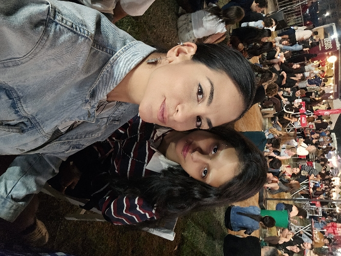 Two women pose for a selfie at an outdoor event. The woman on the top wears a denim jacket. The second woman has dark hair and wears a striped shirt. A large crowd of people sits at tables in the background, illuminated by string lights and various light sources. The event appears lively and well-attended.