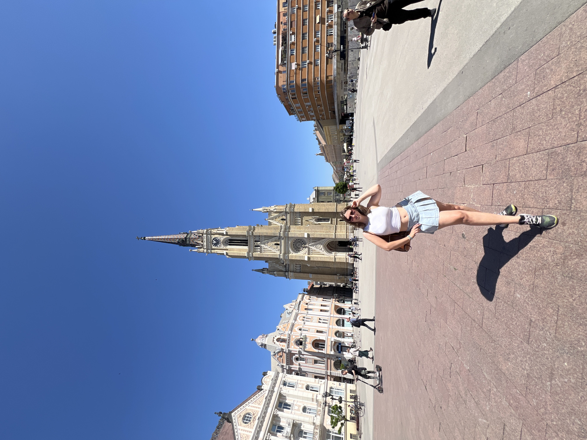 A full-length photograph taken outdoors in Novi Sad, Serbia, features a young woman standing on a brick-paved pedestrian area on a sunny day. She wears a white crop top, a light blue pleated skirt, and dark athletic shoes. Her right arm is raised, hand behind her head. She is standing on the city's central square known as Trg Slobode (Freedom Square). Behind her is the tall spire of the Name of Mary Church, a prominent Roman Catholic church in the square. Further along the square, there are ornate buildings with architectural details. Other people are visible in the background, some walking around the square. Beyond the square are buildings with windows. The sky is bright blue with a few small birds visible in the upper left.