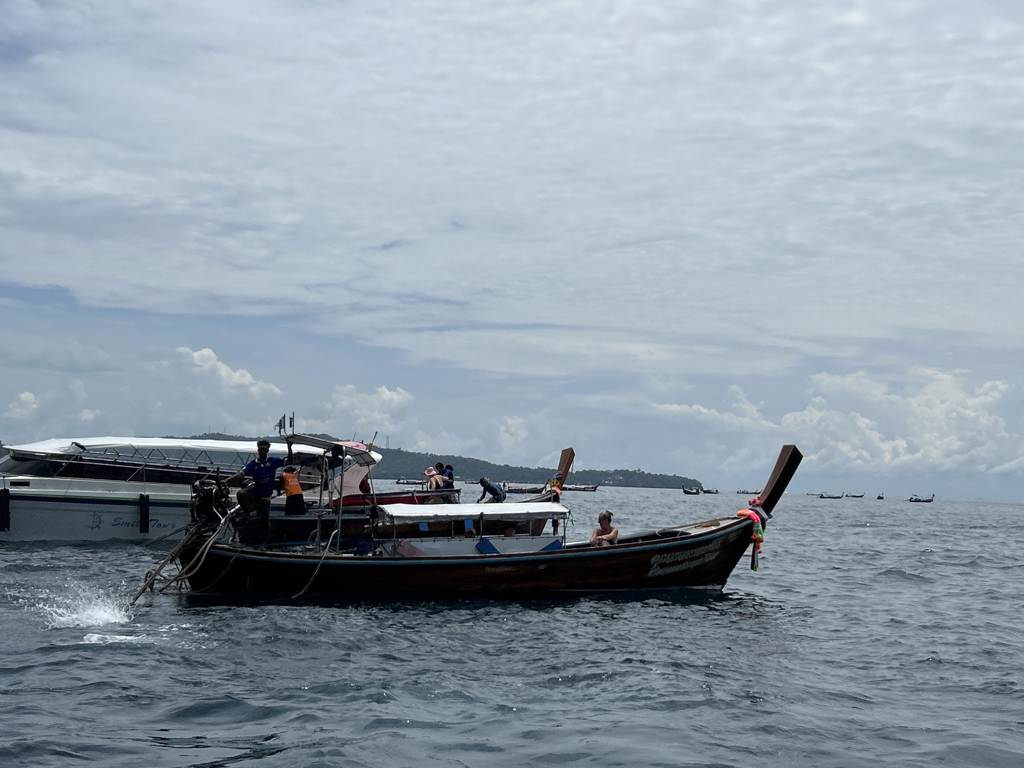 A longtail boat with several passengers is moving through choppy, dark blue water under a cloudy sky. The boat is made of wood and has a distinctive long, pointed prow decorated with colorful tassels. A person is visible at the helm, operating the engine, which is creating a splash of white water. In the background, other longtail boats and a larger, white tourist boat are visible, along with a lush green island in the distance. The scene appears to be in the waters off Ao Nang, Thailand, a popular tourist destination known for its limestone cliffs and beautiful beaches.