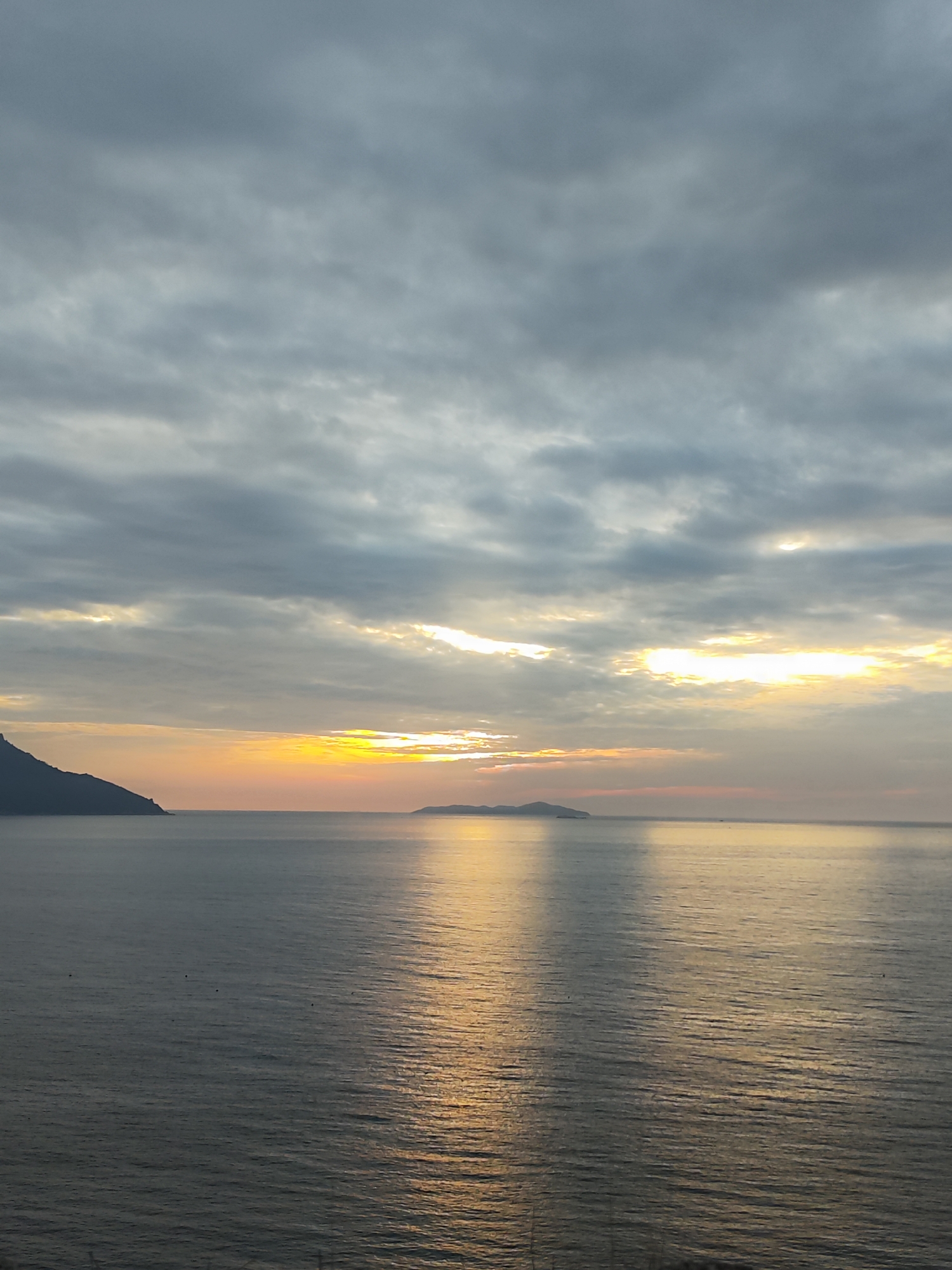 The image depicts a serene seascape at sunrise, likely from a vantage point overlooking the coast of Nha Trang, Vietnam. The sky is overcast with dramatic grey clouds, but a bright band of sunlight pierces through the clouds near the horizon, casting a warm golden glow on the water. The sea is calm, with gentle ripples reflecting the colors of the sky. In the distance, two islands are visible, silhouetted against the dawn light. On the left side of the frame, a dark, mountainous coastline juts out into the sea. The overall impression is one of tranquility and natural beauty.