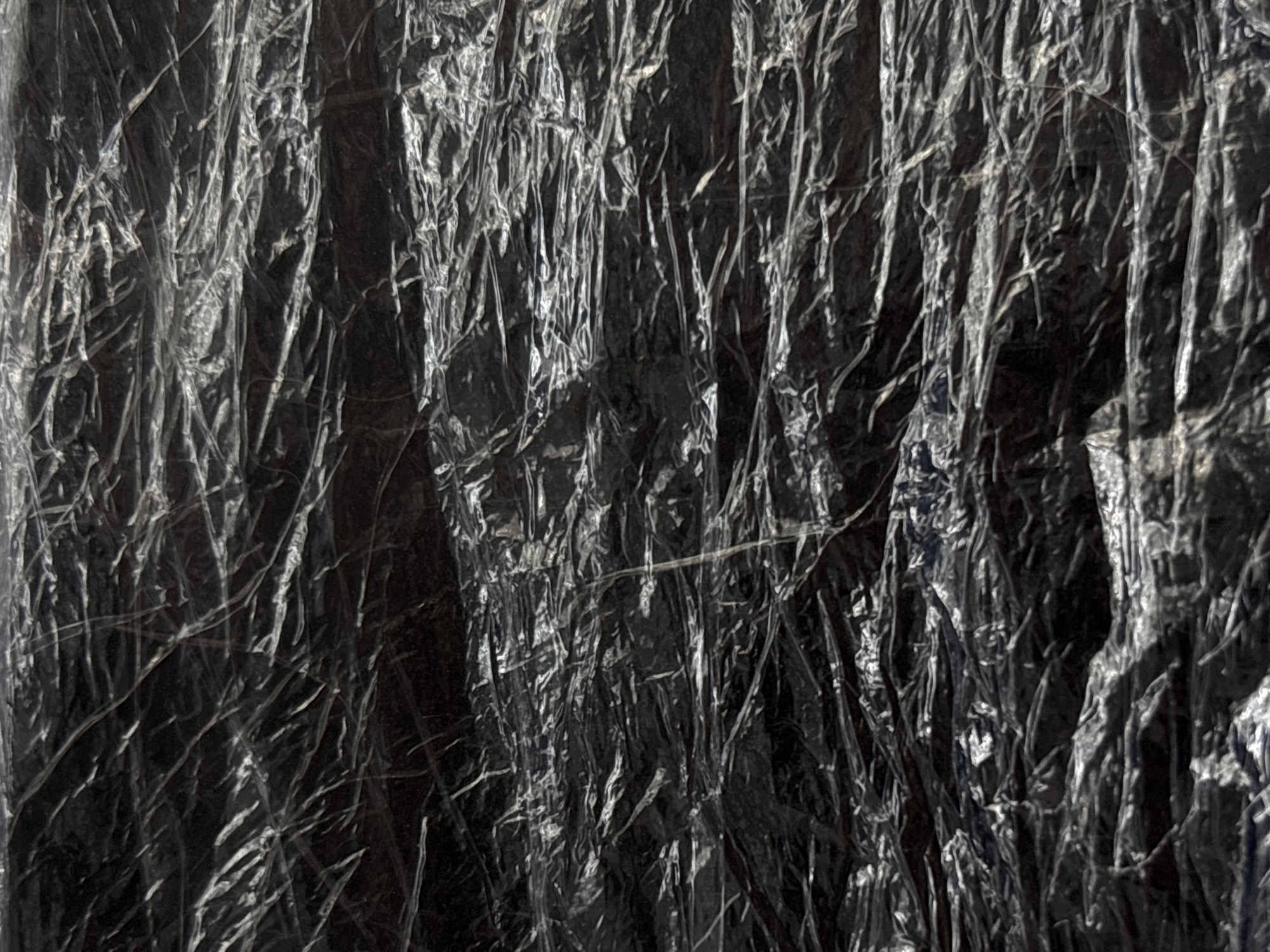 The image is a close-up, abstract shot of a crumpled, dark-colored plastic wrap. The texture is highly detailed, with numerous wrinkles, folds, and highlights from the light source, suggesting it might be stretched over an uneven surface. The plastic appears to be a dark, possibly black or deep gray, material. There are no discernible objects, people, or environmental features visible that would indicate a specific location within Saint Petersburg, Russia, or any other location. The image is purely a study of texture and light on the plastic material.