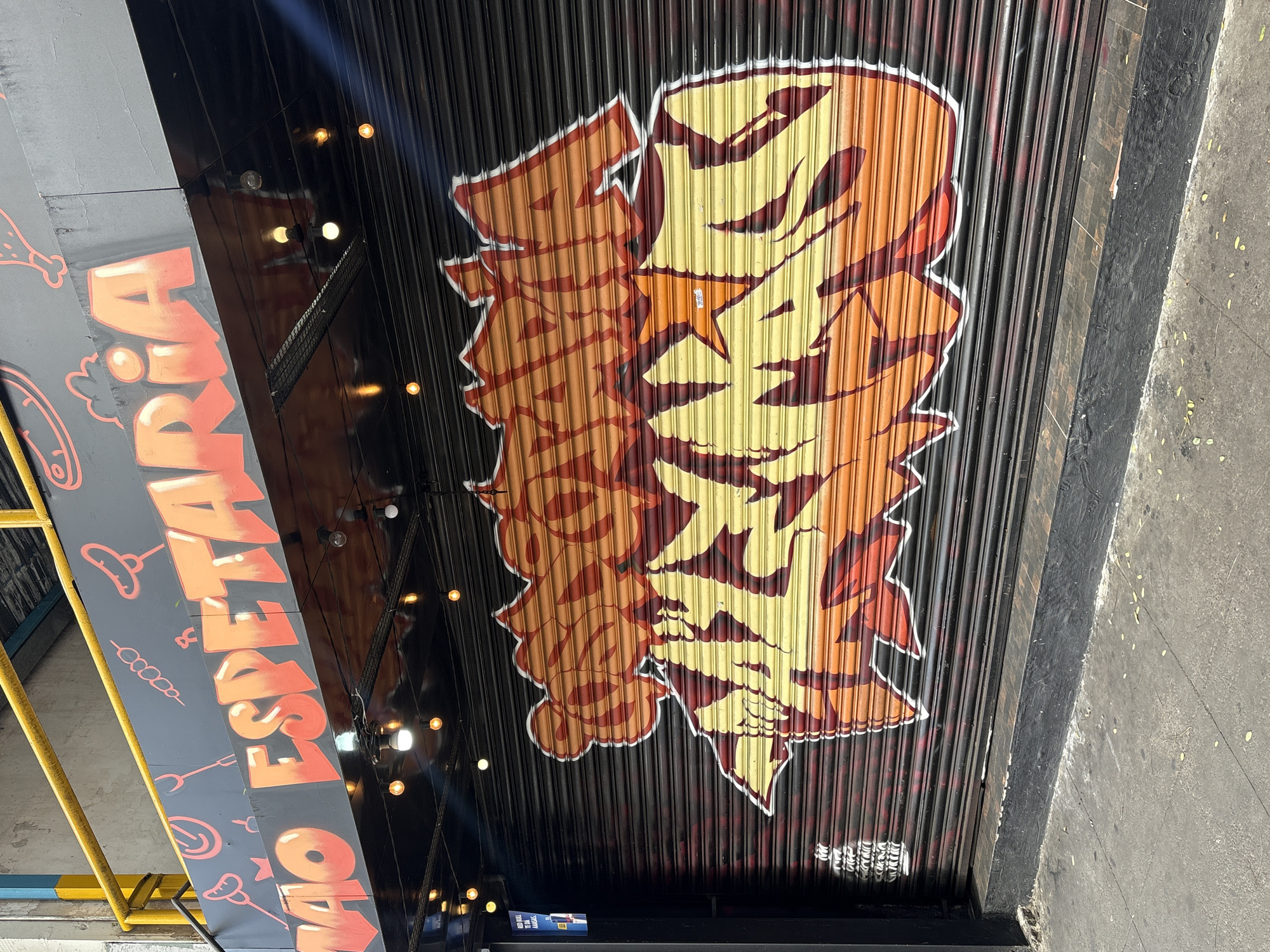 The image shows a closed storefront on a city street in São Paulo, Brazil. The storefront features a black roll-up gate that is heavily graffitied with a large, colorful mural. The mural depicts stylized lettering in shades of orange, yellow, and red, with white and dark red outlines. Above the gate, a black awning displays the words "MAO ESPETARIA" in large, orange, bubble-style letters, also with a red outline. Various red icons, possibly representing food items like sausages and skewers, are scattered across the awning. Small, warm-toned lights are strung across the front of the awning, casting a glow. The street in front of the store is paved with gray concrete, and there is a black painted concrete curb. A small blue sign with white text is affixed to the left side of the building. The overall impression is of a vibrant and artistic urban commercial establishment.
