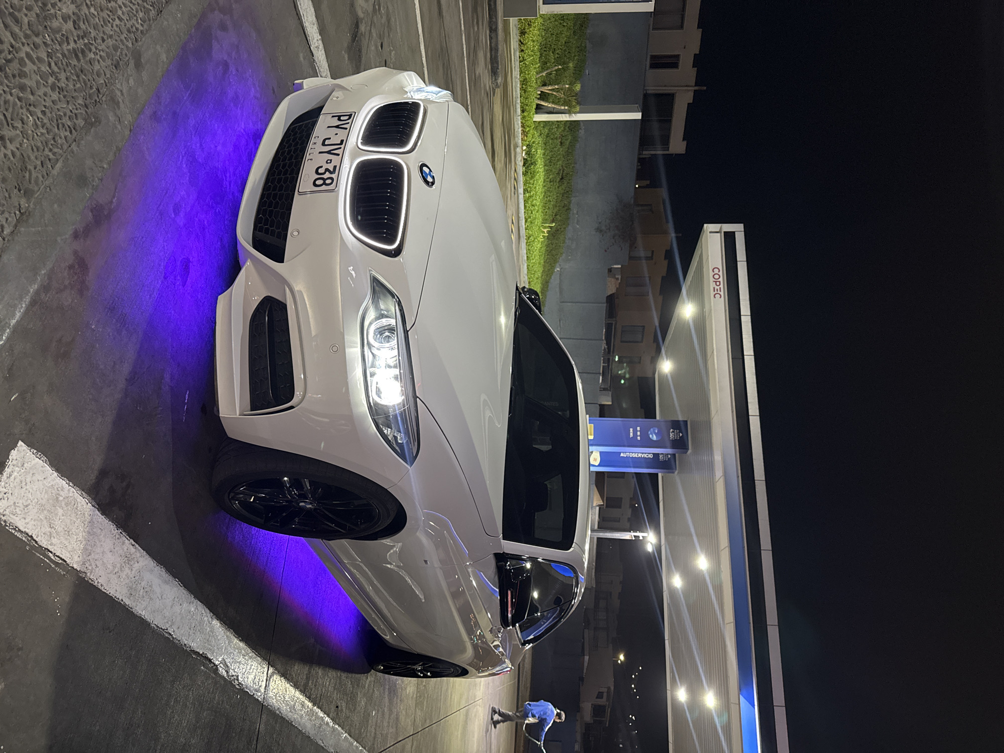 A white BMW is parked at a COPEC gas station in Arica, Chile. The car's license plate reads "PY-JY-38 CHILE". The car is illuminated with blue neon lights. A person wearing a blue jacket stands near the car with an object that appears to be a squeegee. The scene is at night.
