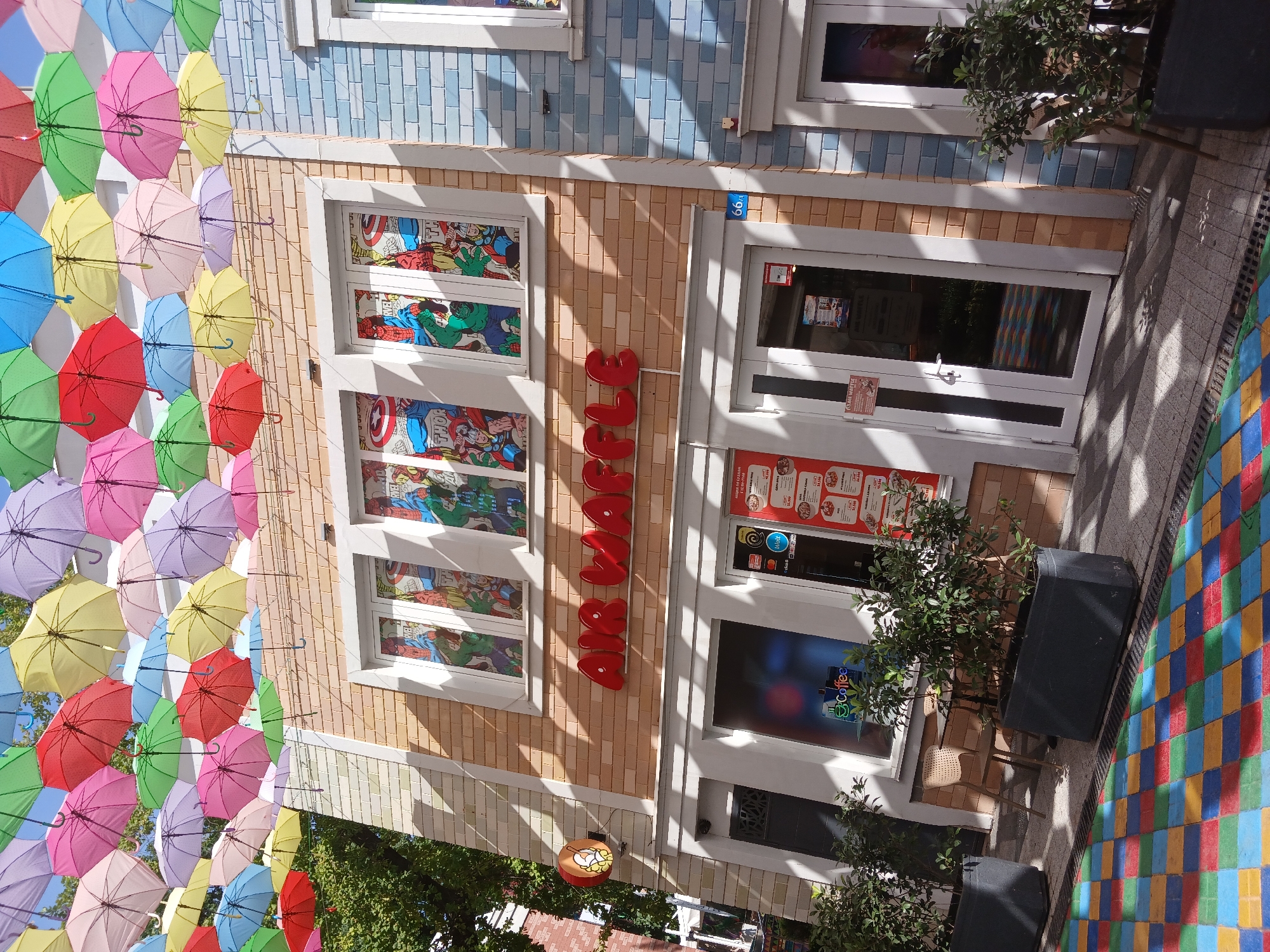 The image shows a brightly decorated building facade, likely a shop or cafe, in Tashkent, Uzbekistan. The building is adorned with an overhead canopy of colorful umbrellas in various shades of pink, yellow, blue, green, and red, creating a vibrant and festive atmosphere. The facade of the building features light brown brickwork on the lower section and light blue tiled accents on the upper part.
There are several windows on the building, some of which are decorated with stained-glass-like panels featuring comic book characters, including Captain America and the Hulk. Large red lettering, spelling out "HANJANG" vertically, is prominently displayed on the brickwork. There are also glass doors leading into the establishment.
Outside the building, there are planters with green trees, and chairs and tables are visible through the glass doors, suggesting an outdoor seating area. The ground in front of the building is paved with a colorful, mosaic-like pattern of red, blue, green, and yellow squares. The lighting and shadows suggest it is daytime, likely sunny. The overall impression is one of a cheerful and welcoming commercial space.