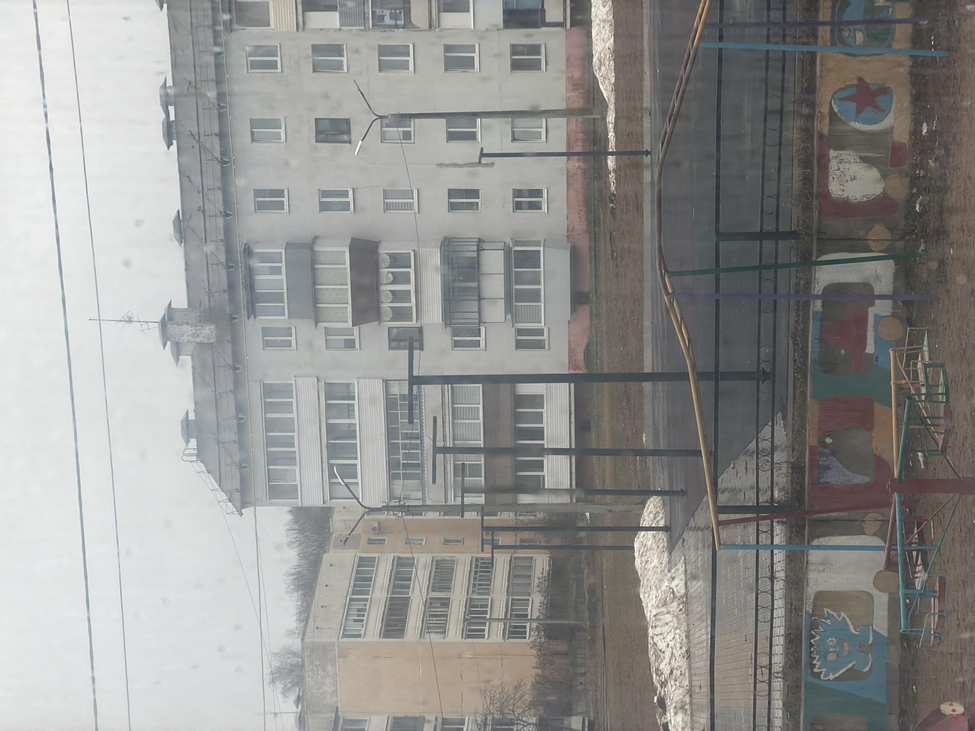 A view of buildings in Noviy Urengoy, Russia, from an elevated vantage point, likely a window. The buildings are multi-story residential structures, predominantly gray in color. Streetlights are present along the sidewalk. A playground is visible in the foreground, with a painted wall featuring a star and an unidentified creature. Remnants of snow are present along the sidewalk. Power lines are visible in the upper-left portion of the frame. The weather appears overcast.