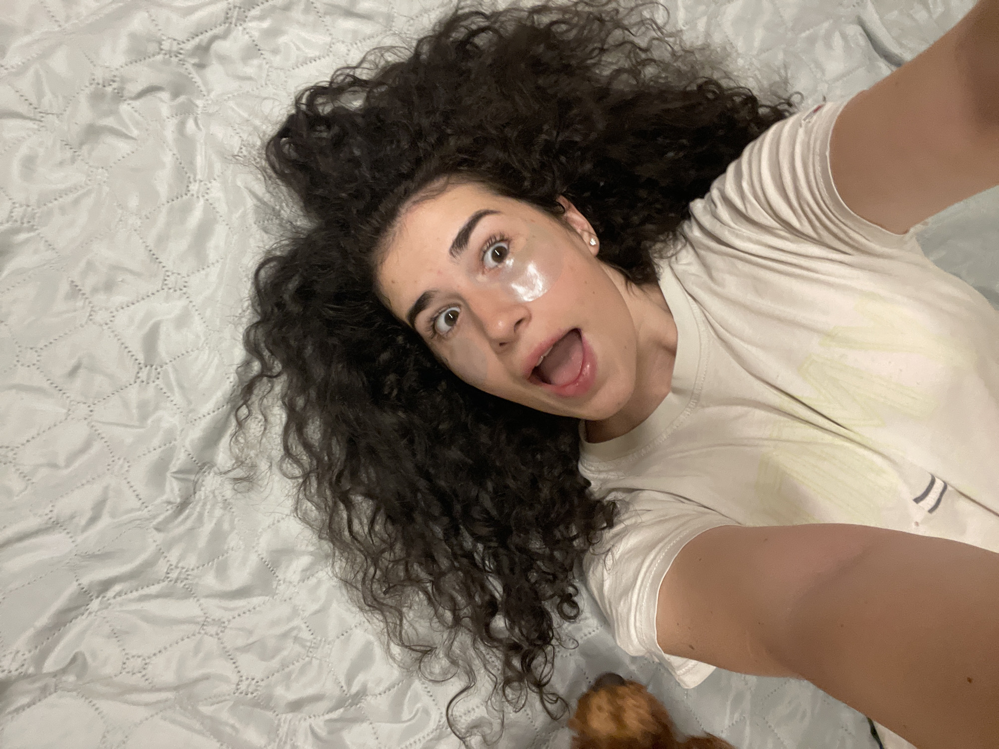 A young woman with long, curly dark hair and under-eye patches lies on a light blue quilted surface, appearing to be a bed or sofa. She is wearing a white t-shirt. Her mouth is open, and her eyes are wide, suggesting a surprised or playful expression. A small part of a brown dog is visible on the left side of the frame. The image is taken from a high angle, looking down at the woman. There are no discernable landmarks or environmental cues that would indicate a specific location within Sofia, Bulgaria.