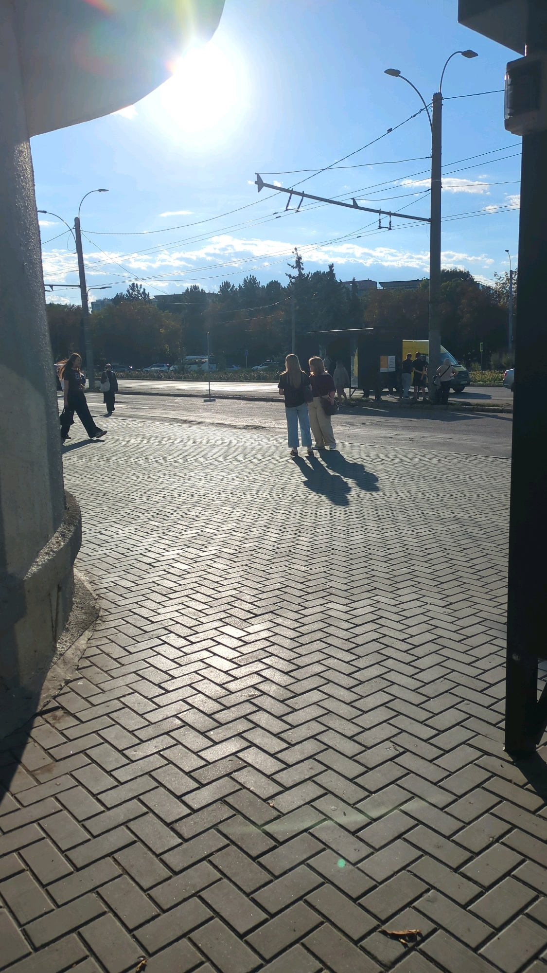 The image depicts a sunlit pedestrian walkway paved with interlocking grey bricks, laid in a herringbone pattern. In the background, a street with traffic and a bus stop can be seen. Overhead, trolleybus wires are strung across the scene, indicating an urban environment. Two young women are walking together on the walkway, their shadows cast long on the brickwork by the low-angled sun. To the left, the edge of a concrete structure is visible. The overall impression is of a typical day in an urban setting, possibly Chișinău, Moldova, given the presence of trolleybus wires and the architectural style.