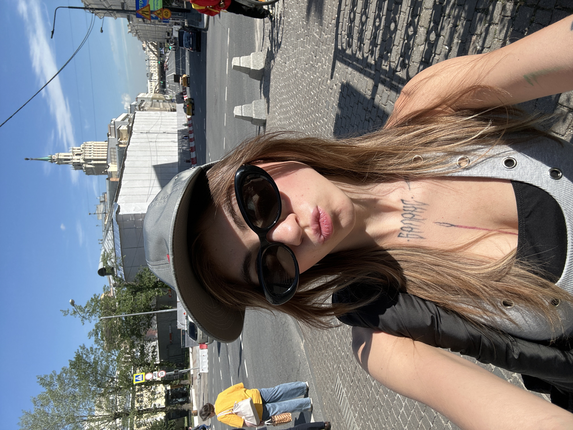 The image is a selfie taken outdoors on a sunny day in Moscow, Russia. In the foreground, a young woman wearing a grey baseball cap and black sunglasses is posing, puckering her lips. She has long, light brown hair and a tattoo on her chest. She is wearing a grey tank top over a black bra. In the background, a street scene unfolds. A tall, ornate building with a spire, characteristic of Stalinist architecture, stands prominently in the distance. The street is lined with buildings, some under construction, and a road with moving traffic, including yellow taxis and black cars. Pavement stones form the ground in the foreground, with shadows cast by the sun. Pedestrians are visible on the sidewalk to the left, and a person on a bicycle or motorcycle is partially visible on the right. Traffic signs are also visible along the street.