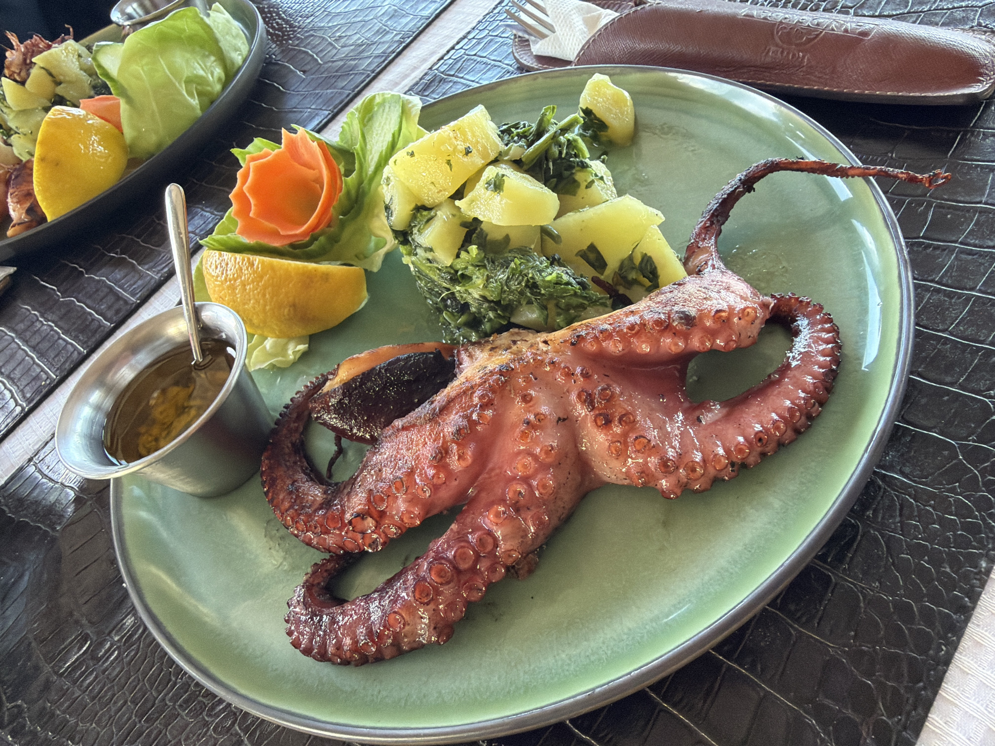 A close-up photograph captures a plate of grilled octopus served at a restaurant in Krašići, Montenegro. The octopus, browned and glistening with oil, is the central focus of the image. It is accompanied by a side of boiled potatoes mixed with leafy greens. A half-slice of lemon is present for seasoning. A small metal container holds a dipping sauce, likely olive oil or a similar vinaigrette. The food is arranged on a light green plate with a metallic rim, set atop a dark brown, textured placemat, which possibly mimics the look of crocodile leather. Another plate containing lettuce and lemon can be seen in the background. A cutlery case lays to the right and a cutlery on the top left of the image.