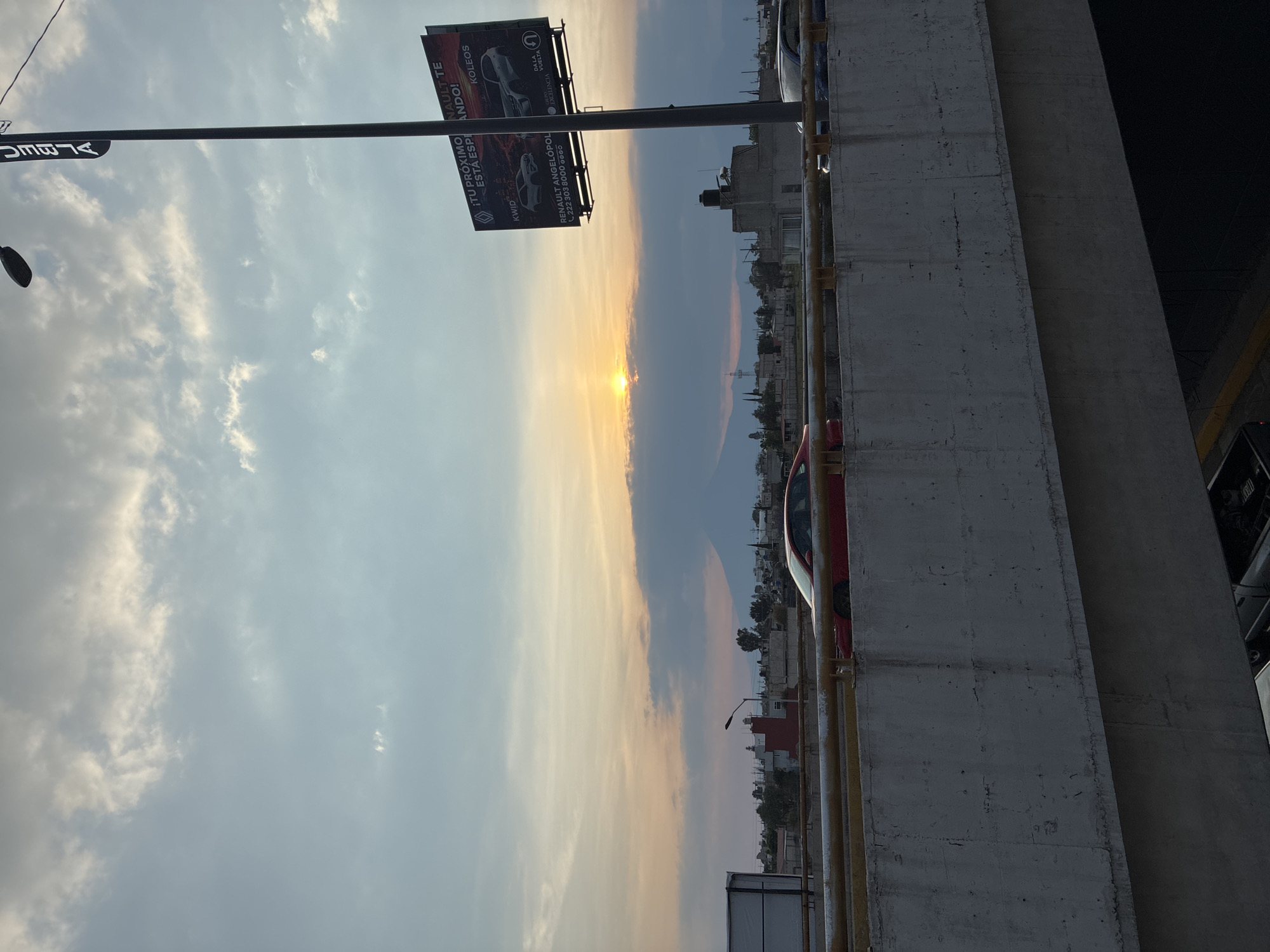 The image shows a sunset scene in San Bernardino Tlaxcalancingo, Mexico, as viewed from a bridge or elevated roadway. A concrete barrier runs along the right edge of the frame, partially obscuring the view. A red car is visible parked next to the barrier. Further into the distance, buildings and trees make up the urban landscape. The sky is filled with clouds, with the setting sun creating a bright, golden hue on the horizon. On the left, a street light and a billboard displaying Renault car models are prominent. The billboard includes the text "¡TU PRÓXIMO ESTA ESPERANDO!". The image is taken during sunset, casting a golden and orange light across the sky and cityscape.