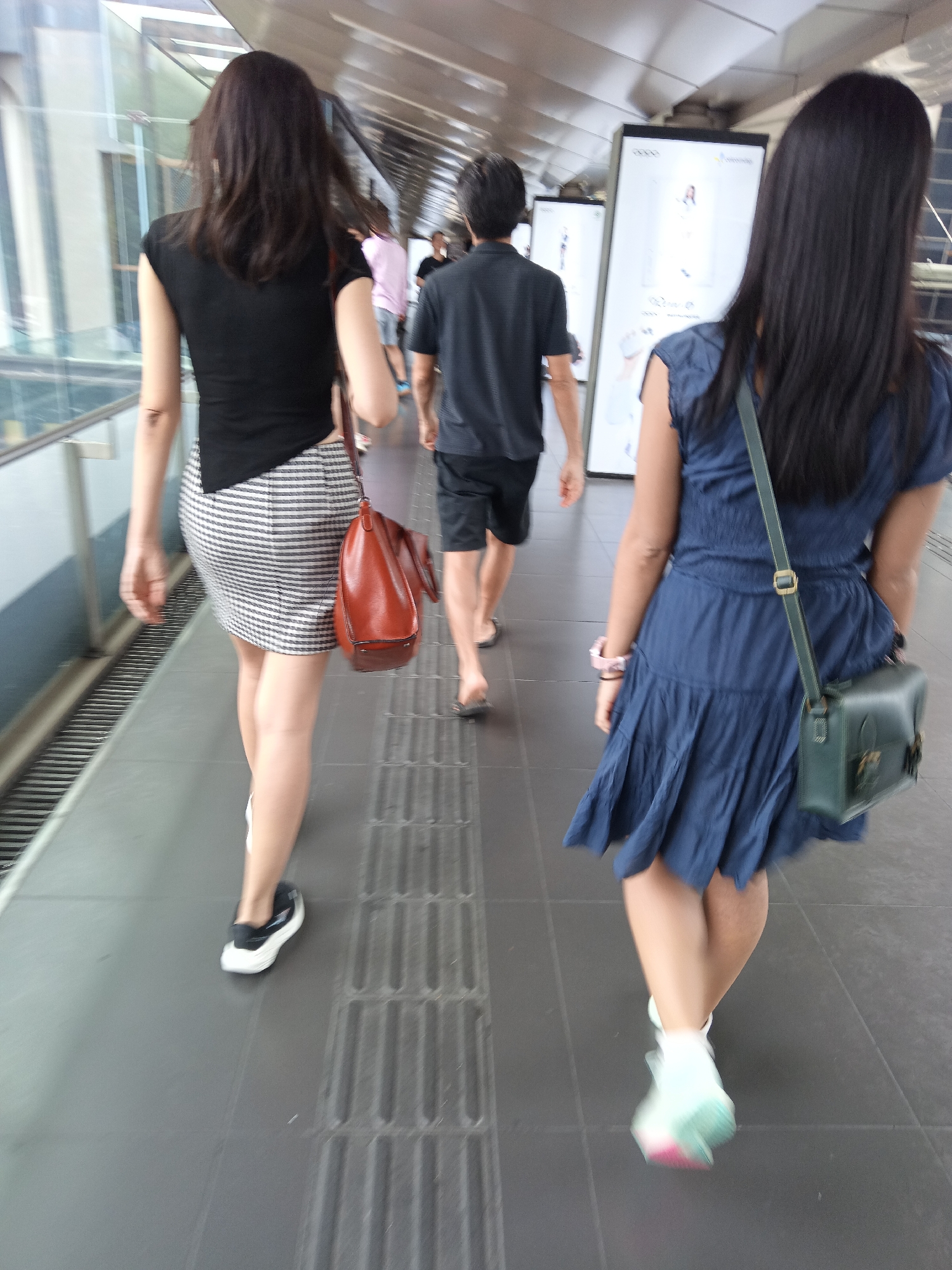 Two women and a man are walking on a covered pedestrian walkway in Kuala Lumpur, Malaysia. The woman on the left is wearing a black sleeveless top and a black and white striped skirt, and is carrying a brown leather handbag. The woman on the right is wearing a blue dress and a green crossbody bag. The man in the middle is wearing a dark t-shirt and black shorts. The walkway has a metal roof and glass panels on the left side, with advertising screens visible in the background. The floor is tiled and has tactile paving. The image appears to be taken during daylight.