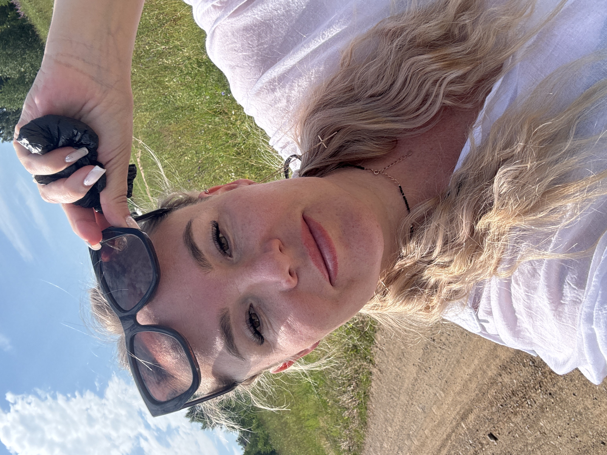 A woman is taking a selfie outdoors on a sunny day. She is wearing a white shirt and sunglasses, which she is holding up with her right hand. Her left hand is holding a black plastic bag. The background consists of a grassy field with some trees in the distance. A dirt path is visible in the foreground. The woman has blonde hair and is wearing makeup. There is a cross-shaped earring and a necklace visible. The sky is blue with white clouds.