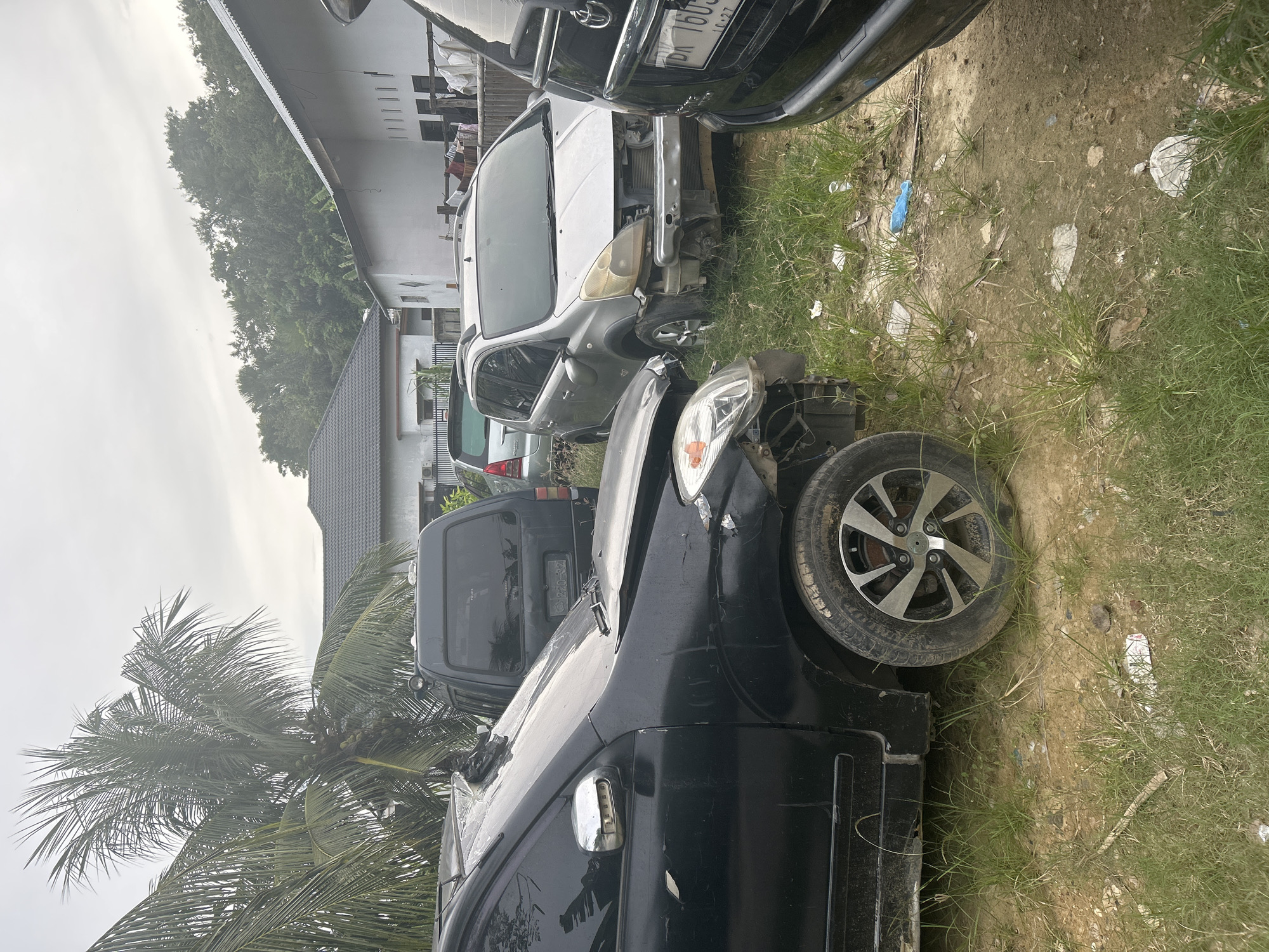The scene shows a car junkyard in Lhokseumawe, Indonesia. Several damaged vehicles are parked on a dirt and grassy lot. The foreground features a black car with front-end damage and a deformed tire. Behind it are a dark-colored van and a silver car with its front bumper missing. A white building with visible laundry hanging on a line and a residential building with a gray roof are in the background. A palm tree is partially visible on the left. The sky is overcast.