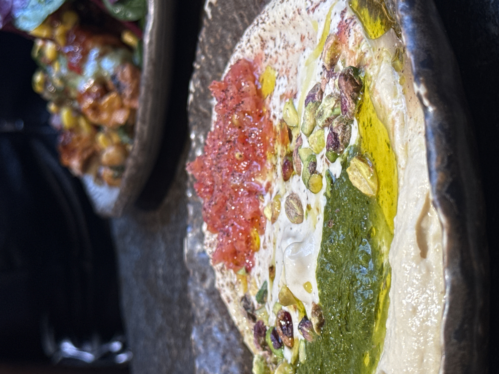 The image captures a close-up, angled view of a vibrant hummus dish served in a dark, rustic bowl or plate. The creamy white hummus is generously topped with an array of colorful ingredients. To the left, a mound of finely chopped red component, possibly tomatoes or roasted red peppers, is visible, dusted with dark spices. A significant portion of the hummus is covered with a rich green paste or sauce, likely a pesto or herb purée, adorned with whole and chopped pistachios that exhibit shades of green and purple. A glistening drizzle of golden olive oil enhances the presentation across the toppings and hummus.
In the softly blurred background, another dish is partially visible, featuring golden-orange, chunky ingredients, possibly grilled corn or a similar preparation. The foreground dish is sharply in focus, emphasizing its textures and brilliant colors under what appears to be soft indoor lighting. No people or text are visible in the frame. The scene depicts a carefully presented meal, possibly in a dining establishment in Barcelona, Spain.
