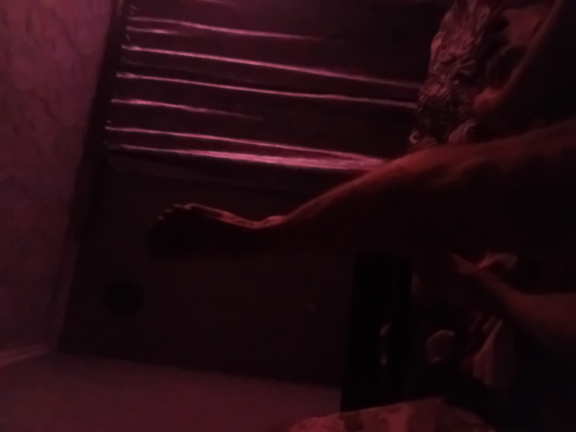 The image depicts a dimly lit bedroom interior, potentially in Verkhniachka, Ukraine, under artificial red lighting. In the foreground, a person's leg is visible, extending from the bottom left of the frame upwards and to the right. The person appears to be lying down or reclining. Behind the leg, a patterned fabric, possibly bedding or a blanket, is visible. Further in the background, draped curtains in a dark color with subtle vertical folds are present on the right side of the frame. The overall ambiance is dark and suggestive, with limited clear details due to the low light conditions and color cast.