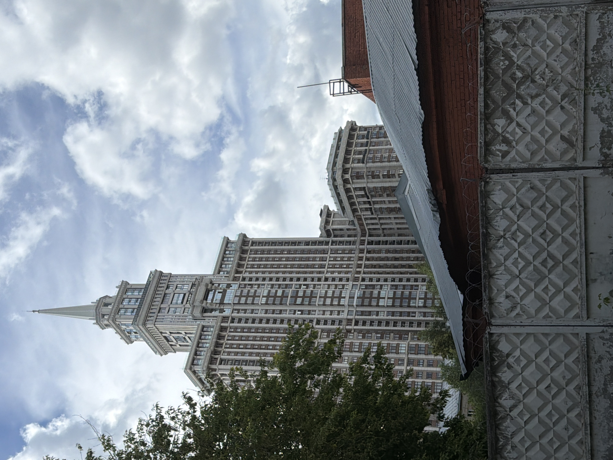 The media file is a photograph taken from a low angle, looking up at a tall, ornate building with a pointed spire. The building is made of stone and has many windows. It is surrounded by trees, and there is a cloudy sky in the background. To the right of the building, a corrugated metal roof and a brick wall are visible. The wall has a pattern of raised diamonds and appears to be old and weathered. There are also strands of barbed wire running along the top of the wall. The overall impression is one of an urban environment, possibly with some industrial elements. The time of day appears to be daytime, and the weather is cloudy. There is no text visible in the image.