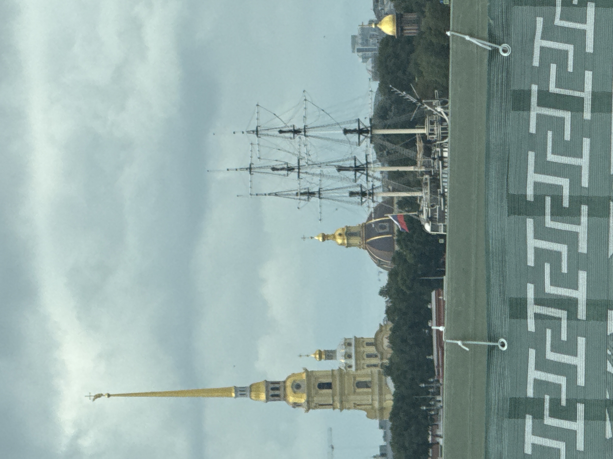 The image depicts a cloudy day in Saint Petersburg, Russia, with a view of the Peter and Paul Fortress. In the foreground, a green mesh barrier with a repeating geometric pattern obstructs the lower portion of the scene. Behind the barrier, a historic sailing ship with three masts is prominently featured. Further in the background, the iconic golden spire of the Peter and Paul Fortress Cathedral pierces the sky, accompanied by other church domes. Dense green trees line the waterfront, and the cityscape stretches beyond. No people are visible in the image.