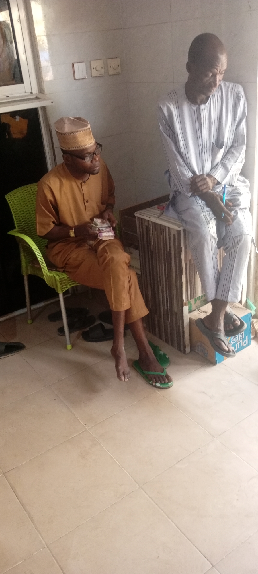 The image shows two men indoors. One man is seated on a green plastic chair wearing a brown traditional outfit and glasses, holding a stack of money. The second man is seated on what appears to be a decorated platform. He is wearing a white and blue striped outfit and slippers, with a pen in his hand. Several pairs of sandals or flip-flops are on the floor near the seated men. The floor is tiled. There are white wall sockets or light switches on the wall. A window with a closed blind is visible. The location is in Sabon Gari West, Nigeria.