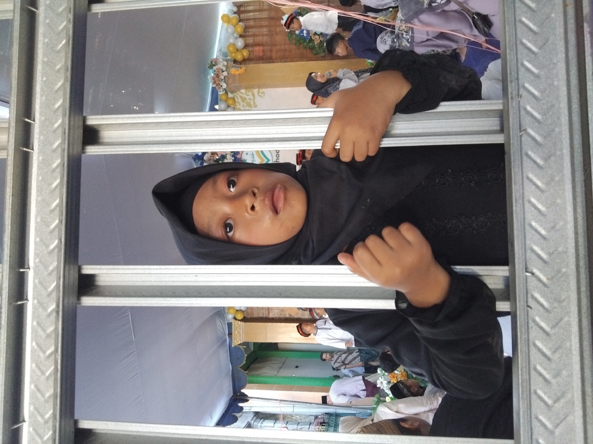 The image shows a young girl wearing a black hijab and black clothing, looking at the camera through metal bars. She is holding onto the bars with her hands. In the background, there are other people, some of whom are also wearing hijabs and traditional clothing. The scene appears to be an outdoor event, possibly a religious gathering, with decorations such as balloons and flowers visible. The environment suggests it is in Kepuh Kiriman, Indonesia, given the cultural context indicated by the clothing and the likely setting for such an event. The lighting suggests it is daytime.