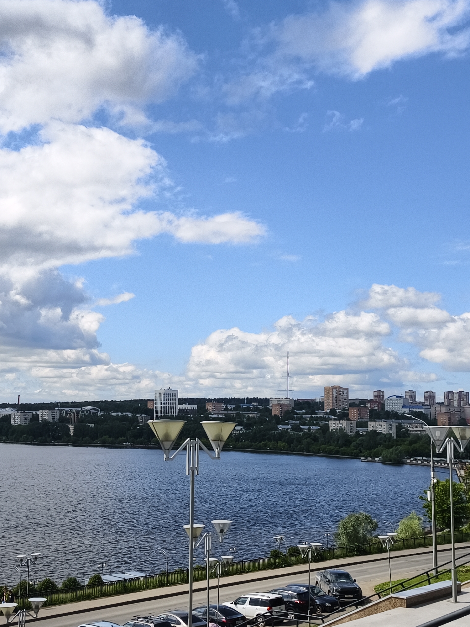 The image presents a wide view of a city situated alongside a large body of water under a partly cloudy sky. The sky above is a bright blue with numerous white cumulus clouds scattered across it, suggesting clear weather.
In the midground, a dark blue lake or wide river stretches horizontally across the frame, its surface rippled by light breezes. On the far bank, a sprawling cityscape unfolds, characterized by a mix of multi-story residential buildings and a few taller, more modern structures, including one distinct white high-rise. Green trees and dense foliage line the water's edge and are interspersed among the buildings, indicating a well-treed urban environment. A prominent tall, slender communications tower with cross-bracing rises above the distant skyline on the left, a recognizable landmark.
The foreground features an elevated paved area, likely a promenade or a road, running parallel to the water. A series of distinctive streetlights with double, inverted-pyramid or conical light fixtures stand along this path, casting reflections on their metal poles. Several cars, including SUVs and sedans, are parked along the side of the road. A metal railing runs along the edge of the elevated area, separating it from what appears to be a lower embankment or walkway nearer the water. On the far right, a concrete structure, possibly a bench or part of a terrace, is visible.
The scene is illuminated by bright daylight, suggesting a morning or early afternoon setting. No people are distinctly visible, and there is no discernible text in the image. The overall impression is that of a serene urban landscape viewed from an elevated perspective on a pleasant day in Izhevsk, Russia.