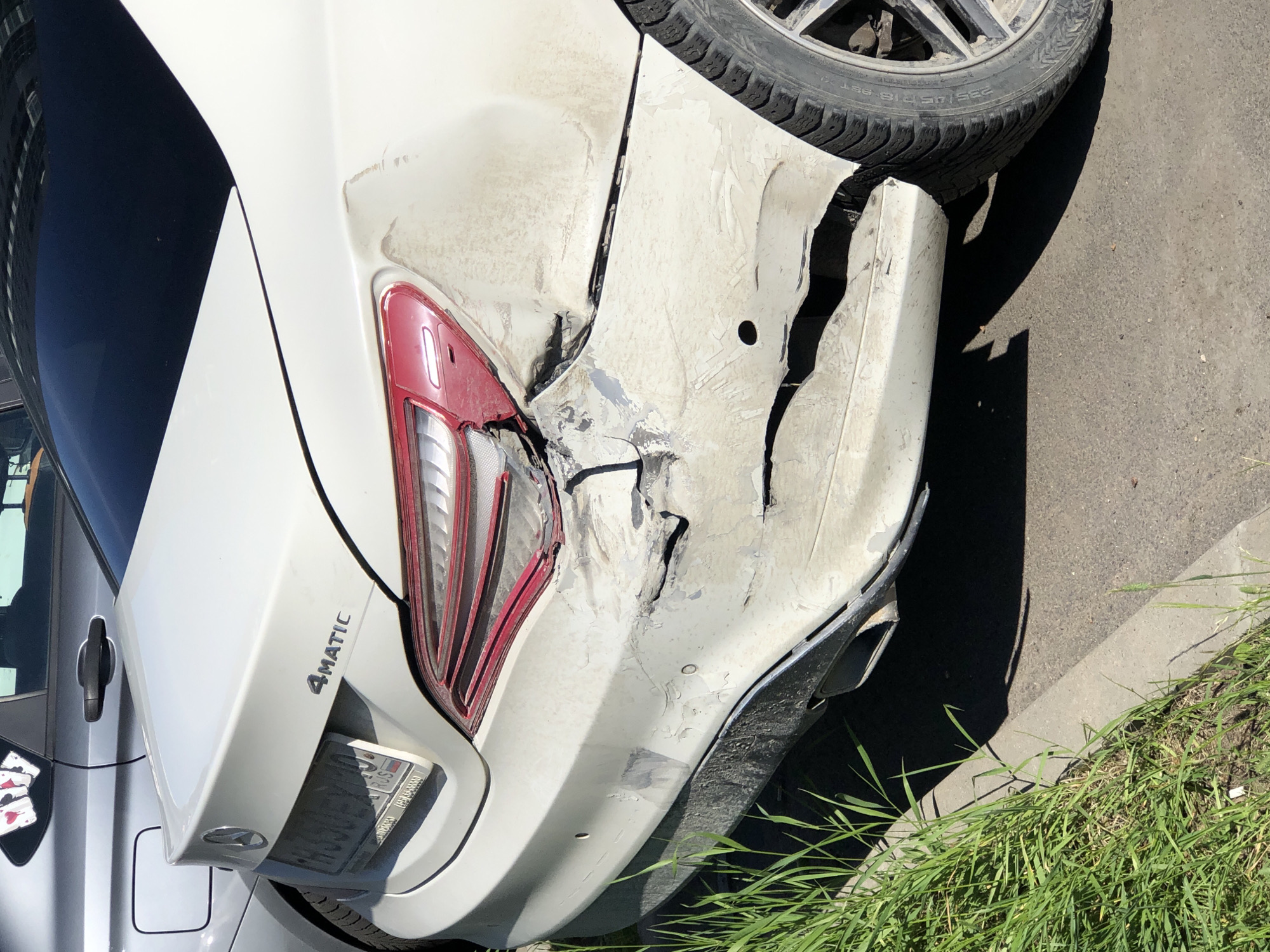 The image shows the rear passenger side of a white Mercedes-Benz CLA-Class sedan with significant damage to the bumper and quarter panel. The car is parked on a paved surface next to a curb, with tall green grass growing on the left side of the image. In the background, another car, a silver sedan, is parked closely to the Mercedes. The Mercedes has a "4MATIC" badge on its trunk. The license plate is visible and appears to be a Russian plate. The damage to the Mercedes is extensive, with large dents, scraped paint, and broken plastic pieces. The taillight on the damaged side is also partially broken. The scene suggests the car has been involved in a collision. The presence of Russian license plates indicates the location is likely in Russia.