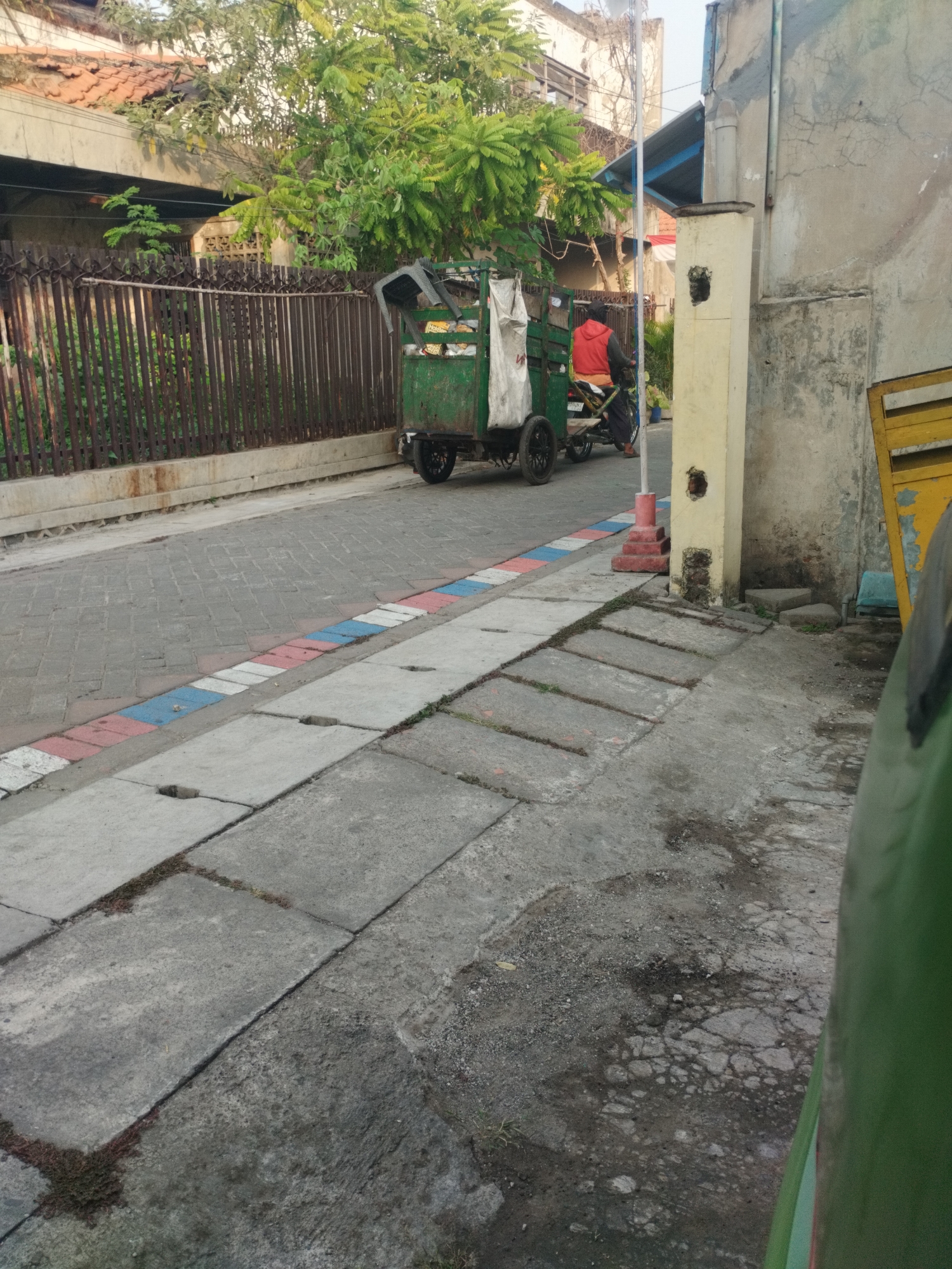 The image depicts a street scene in Surabaya, Indonesia. In the background, a person wearing a red jacket rides a motorcycle pulling a green cart. The cart appears to be a type of mobile vendor or collection vehicle. The street is paved with cobblestones, bordered by a sidewalk. The sidewalk has a decorative stripe of red, white, and blue tiles. To the right of the street, a light yellow pillar stands next to a textured gray wall. Further to the right, a portion of a yellow structure is visible. Trees and buildings are present in the background, suggesting an urban or semi-urban environment. The overall lighting suggests it is daytime, possibly late afternoon due to the shadows.