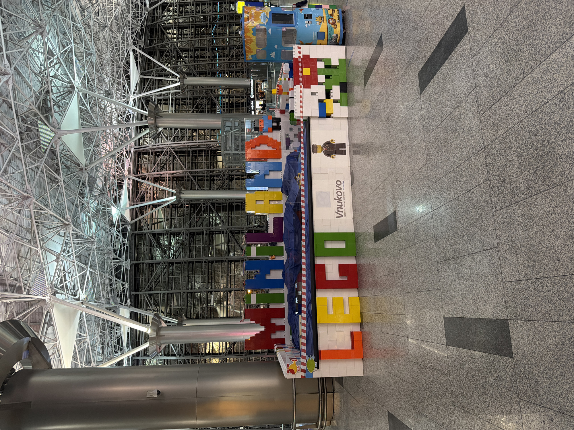 The image shows a large, colorful LEGO-themed display set up inside an airport terminal. The display features oversized LEGO bricks forming the words "MIL" and "LAND" in large, blocky letters, along with the word "LEGO" in prominent, separate bricks on a white base. A small LEGO-style figure of a man in a pilot's uniform stands to the right of the "LEGO" spelling. The base of the display has "Vnukovo INTERNATIONAL AIRPORT MOSCOW" printed on it. Behind the display, the architecture of the airport is visible, with large metal pillars and a complex, industrial-style ceiling structure. To the far right, a blue and yellow structure resembling a LEGO bus or vehicle is also part of the display. The floor is tiled with a speckled gray pattern. There are no visible people interacting directly with the display in the foreground. The scene appears to be a public space, likely an airport concourse, in Vnukovo, Russia, given the text on the display.