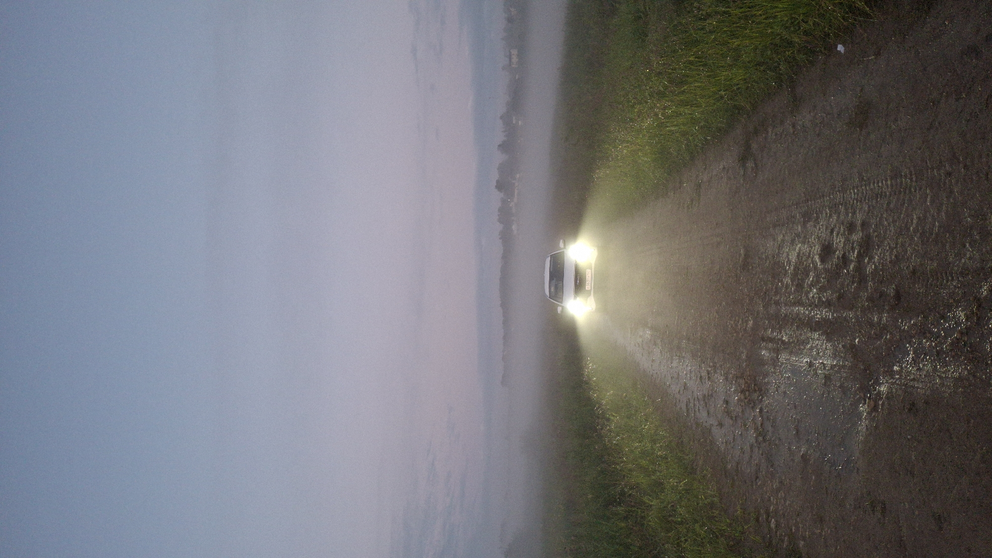 A white car with its headlights on drives down a muddy dirt road in Kaporskoe, Russia, at dusk. The car is facing the camera, and its headlights illuminate the road and the surrounding fog. The road is wet and reflects the headlights. On either side of the road are grassy fields. In the distance, silhouetted trees and buildings can be seen through the fog. The sky is a pale blue and gray, indicating the transition from day to night.