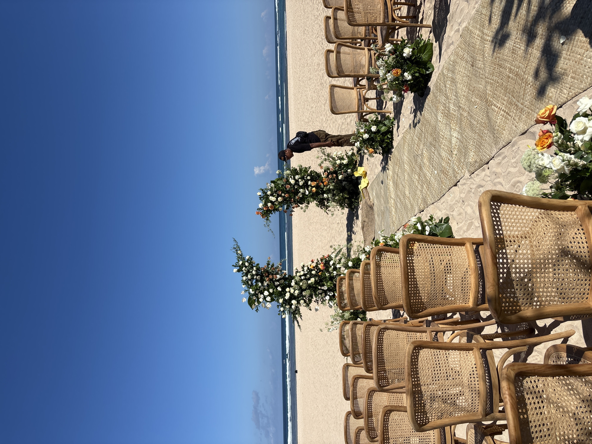 The image captures an outdoor wedding setup on a beach in Nusa Dua, Indonesia. Rows of wooden, rattan-backed chairs are arranged on the sand, facing a floral arch. A woven mat is placed on the sand to create an aisle. A man is positioning flowers on the arch, indicating preparations are underway. The clear blue sky contrasts with the white sand beach and the turquoise ocean.