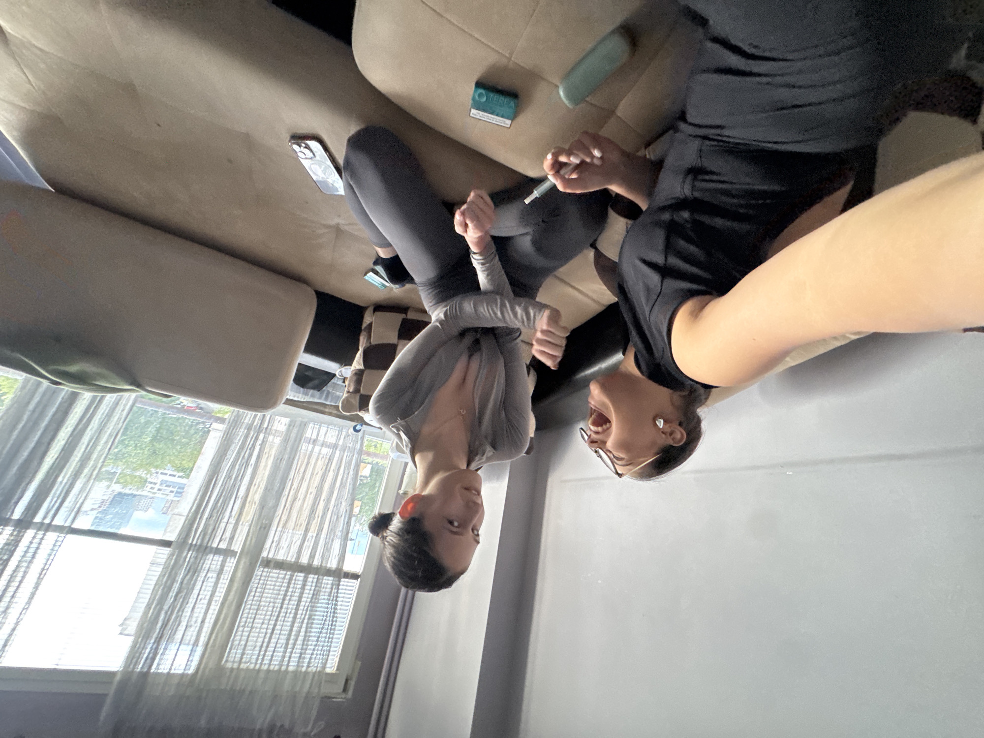 Two women are posing for a photo. One is wearing a gray sweater and black leggings, the other is wearing a black top and shorts. They are sitting on a beige couch. On the couch, there is a black cell phone, a pack of teal-colored cigarettes, and a lighter. Outside the window, buildings can be seen.